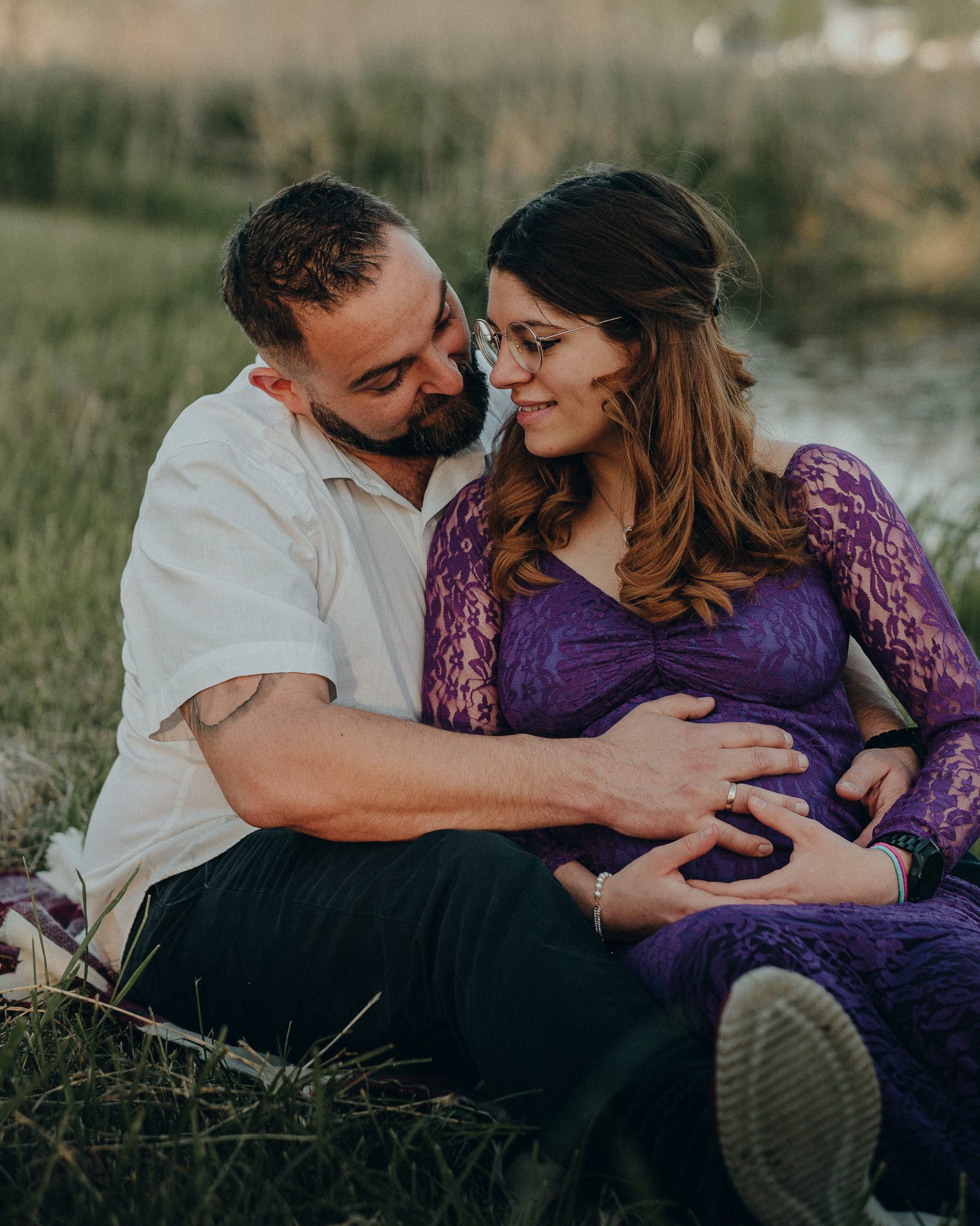 Ein verliebtes Paar sitzt im Freien am Wasser, die Frau ist schwanger, beide lächeln sich liebevoll an, die Frau trägt ein lila Kleid mit Spitzenmuster.