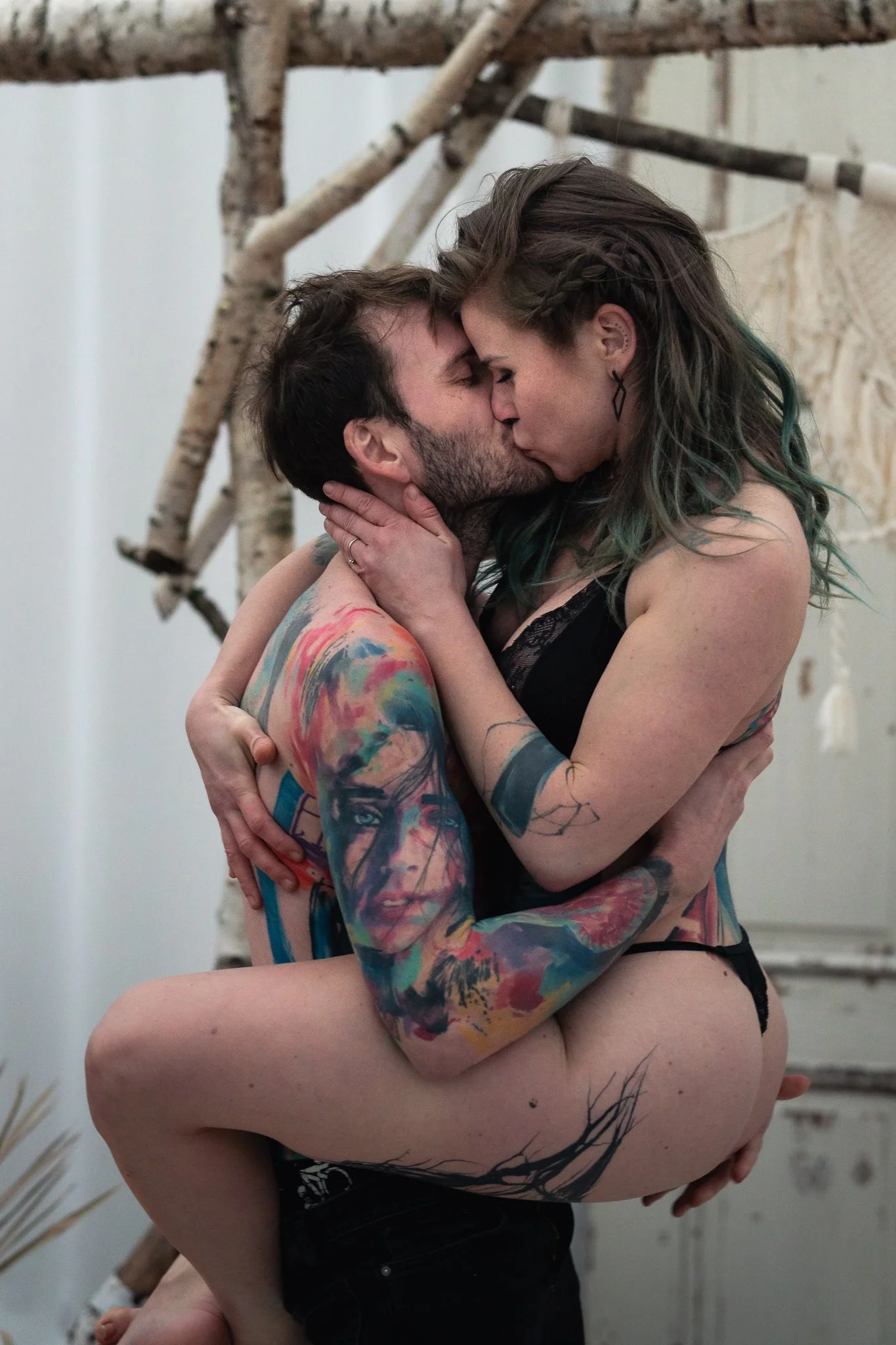 Ein Paar küsst sich innig, der Mann trägt Tattoos und der Frau hat gestreiftes Haar, beide in intimem Moment in einem Raum mit natürlichen Holzdekorationen.