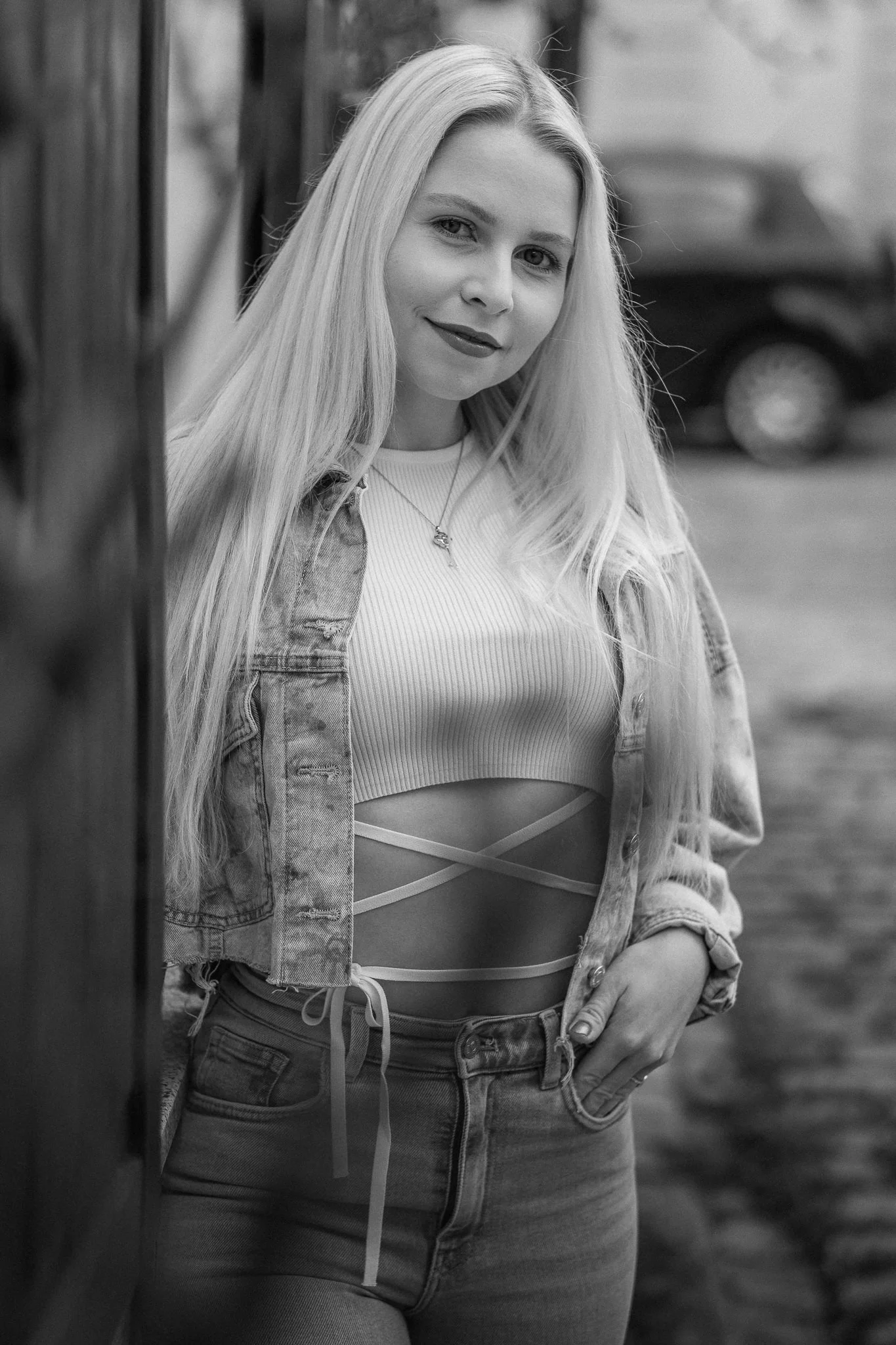 Porträt eines lächelnden jungen Frauen mit langen blonden Haaren, die an einem Holzpfosten in einer Straße steht, im Hintergrund ein Auto