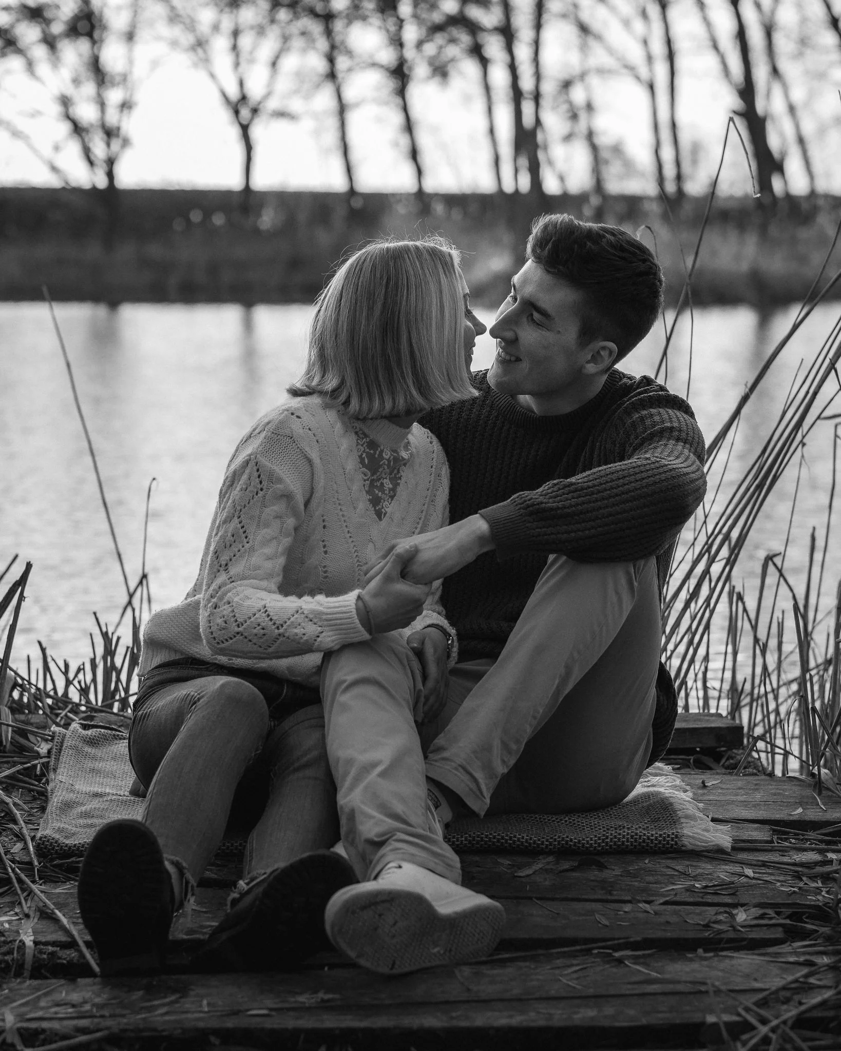 Ein junges Paar sitzt am Ufer eines Sees, umarmt sich und schaut sich liebevoll an, im Hintergrund Bäume, schwarz-weiß Foto.