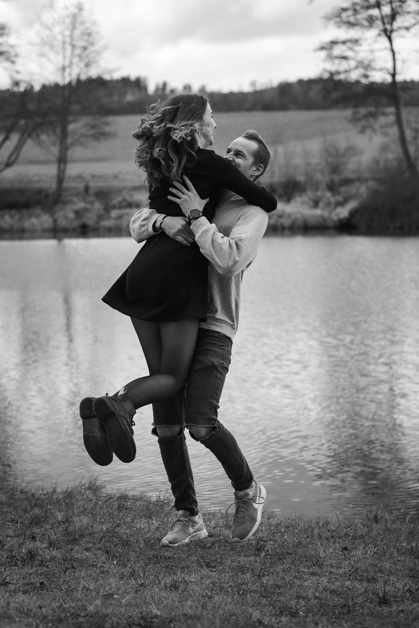 Junge Frau und junger Mann umarmen sich lachend am Seeufer, die Frau wird hochgehoben, im Hintergrund Bäume und Wasser, Schwarz-Weiß-Foto.