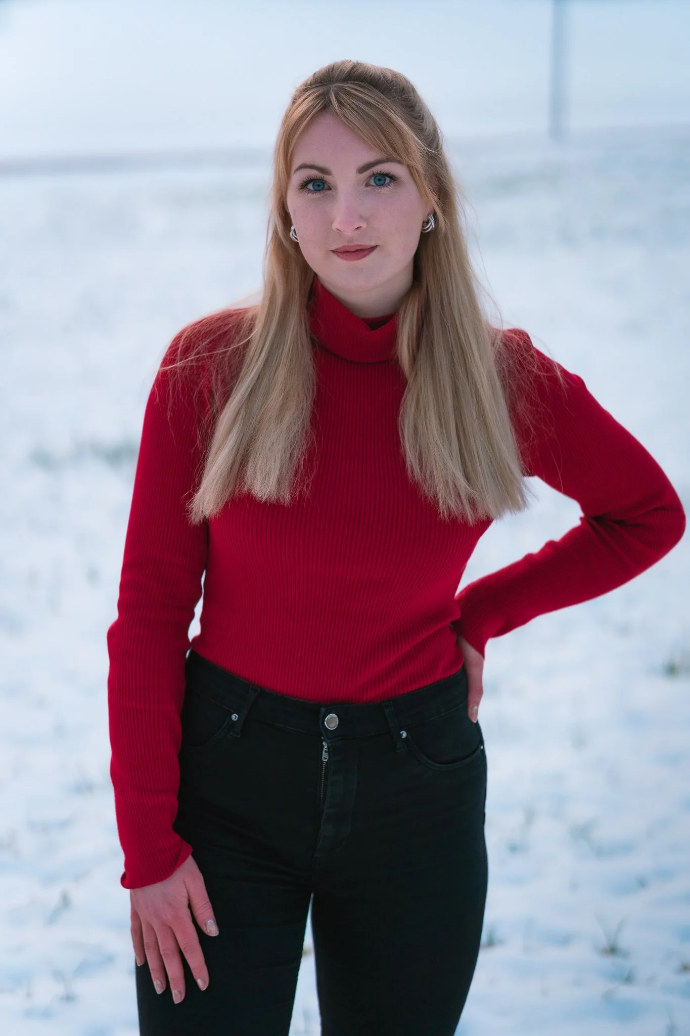 Junge Frau mit blonden Haaren, blauen Augen, rotem Pullover und schwarzen Hosen steht vor verschneitem Hintergrund.