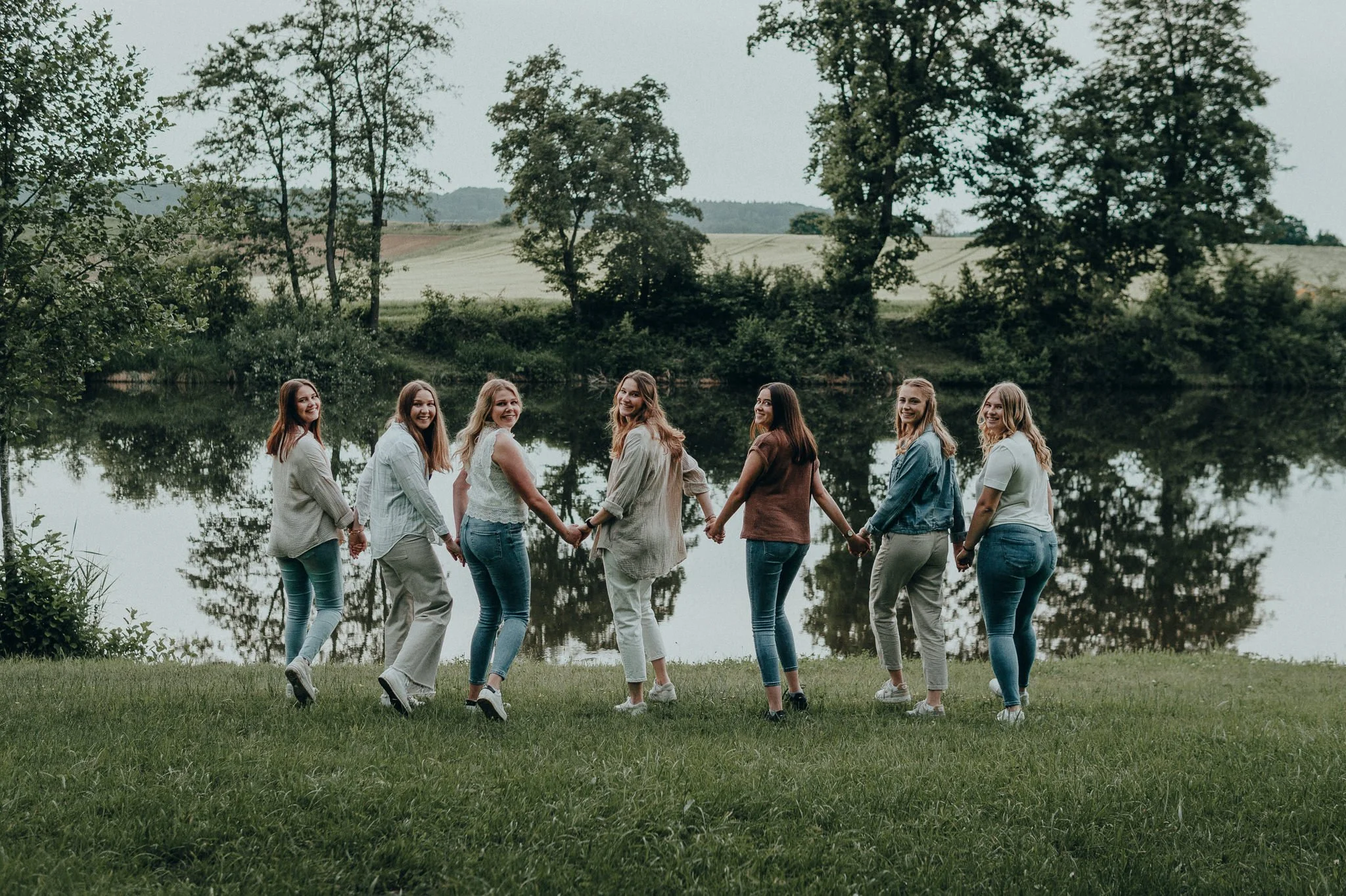 Sieben junge Frauen halten sich an den Händen und lachen am Ufer eines Sees, umgeben von Bäumen und grünen Wiesen.