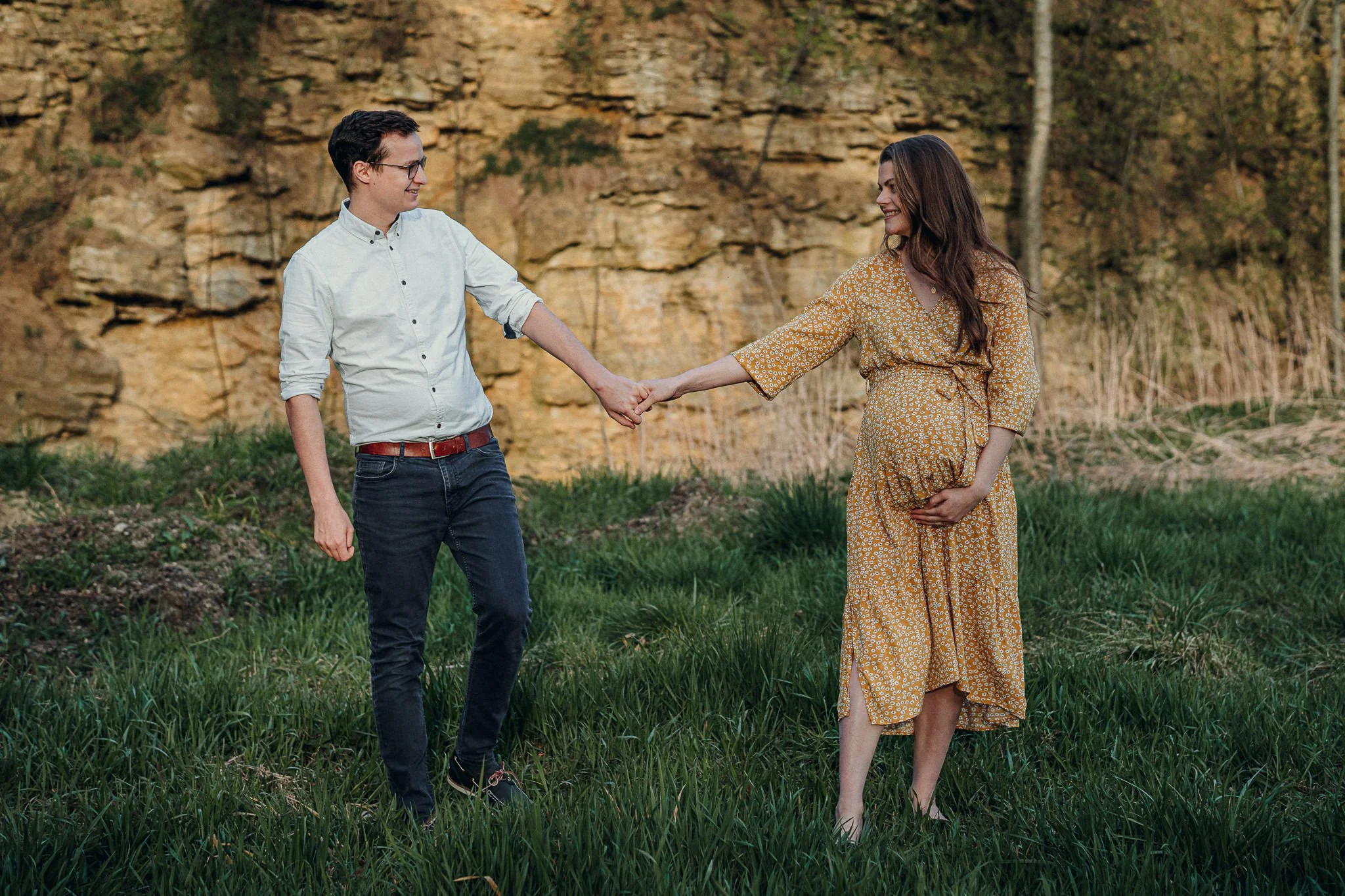Ein Mann und eine schwangere Frau halten sich an den Händen, während sie im Freien auf dem Gras stehen, mit einem Felsen und Bäumen im Hintergrund.