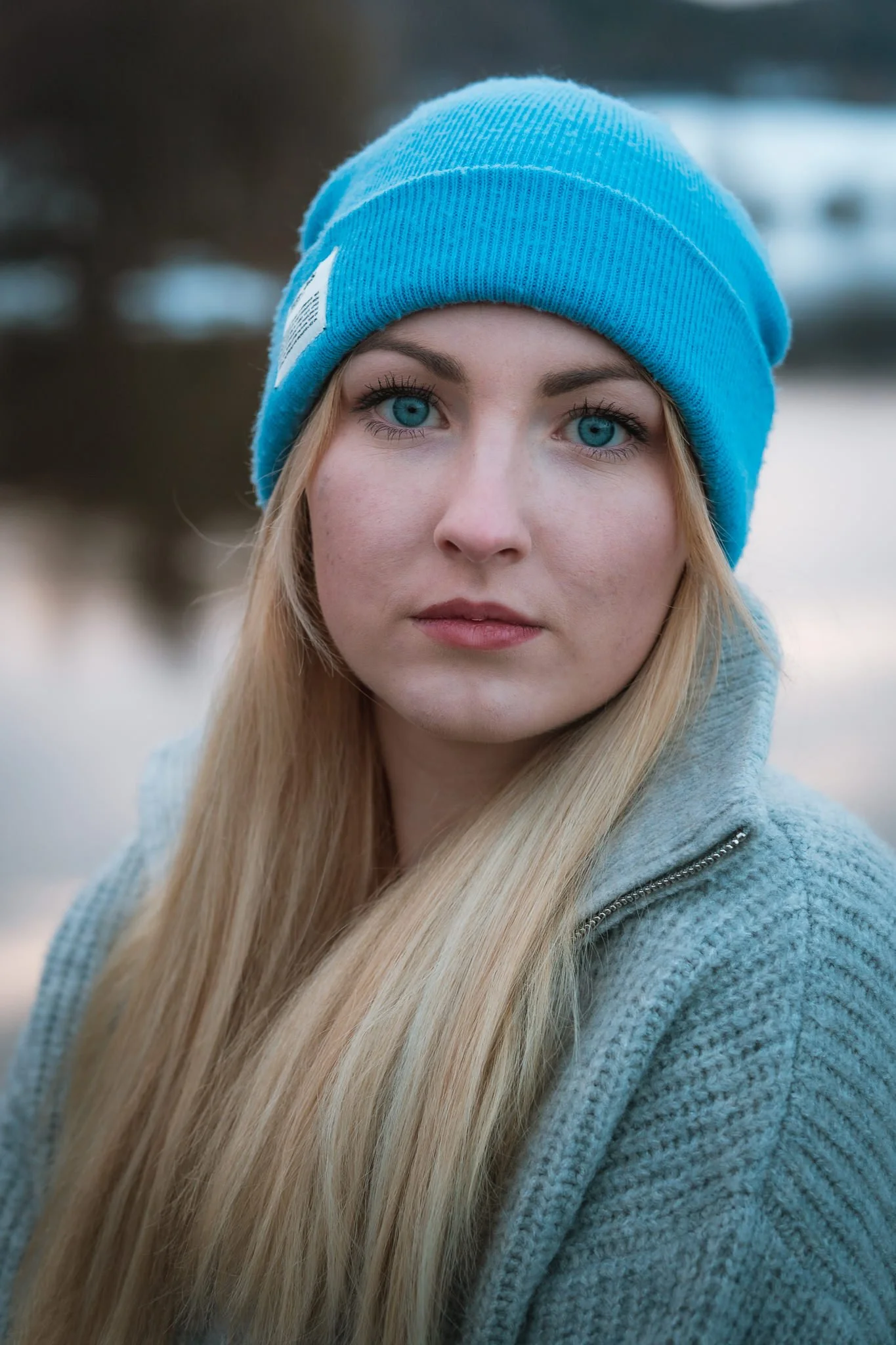 Nahaufnahme eines jungen Mädchens mit blauen Augen und langen blonden Haaren, das eine blaue Strickmütze trägt, im Hintergrund ein Wasser und Bäume.