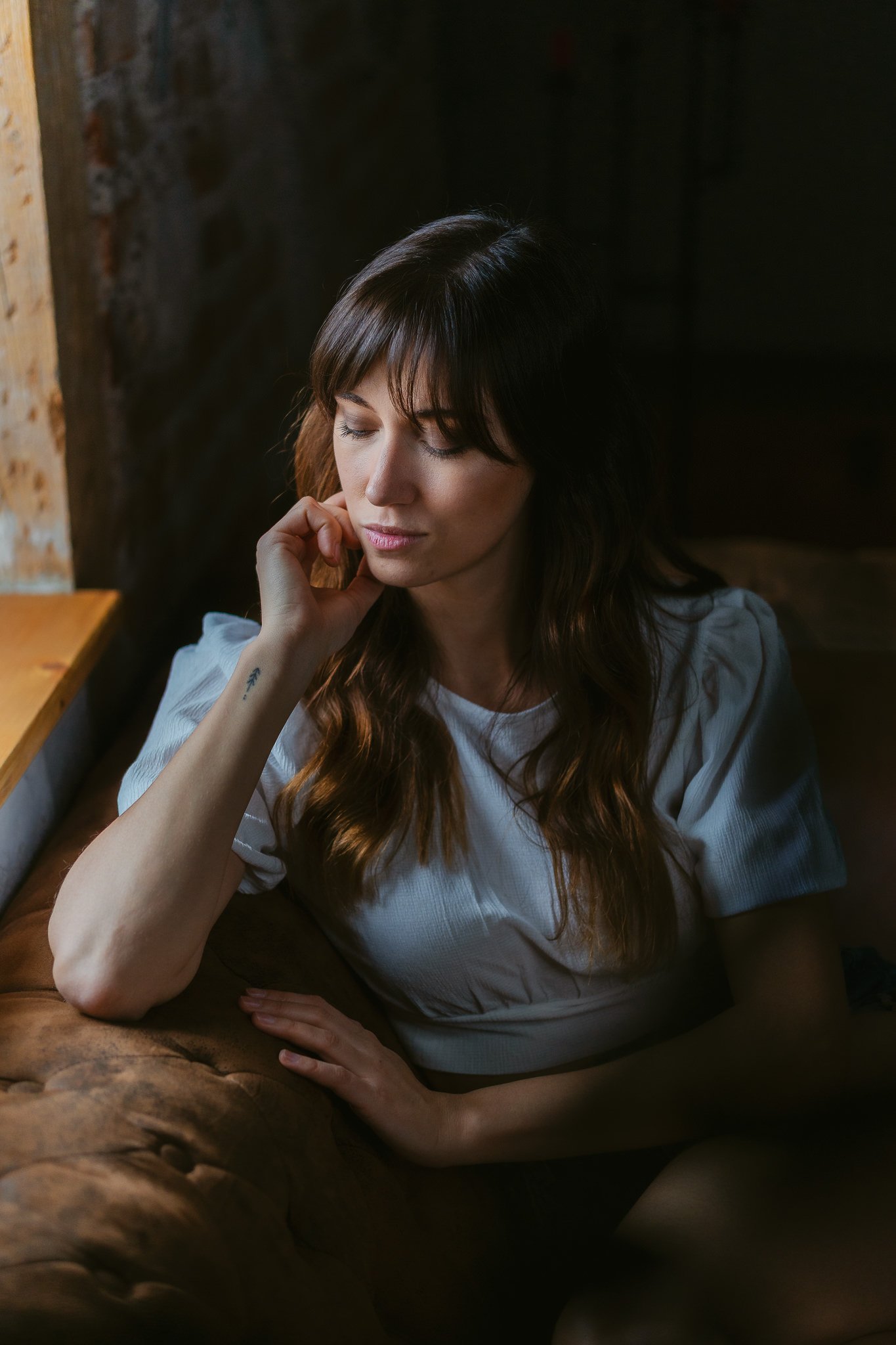 Frau mit braunen Haaren und weißen Oberteil, nachdenklich, sitzend auf braunem Sofa, dunkle Hintergrund.