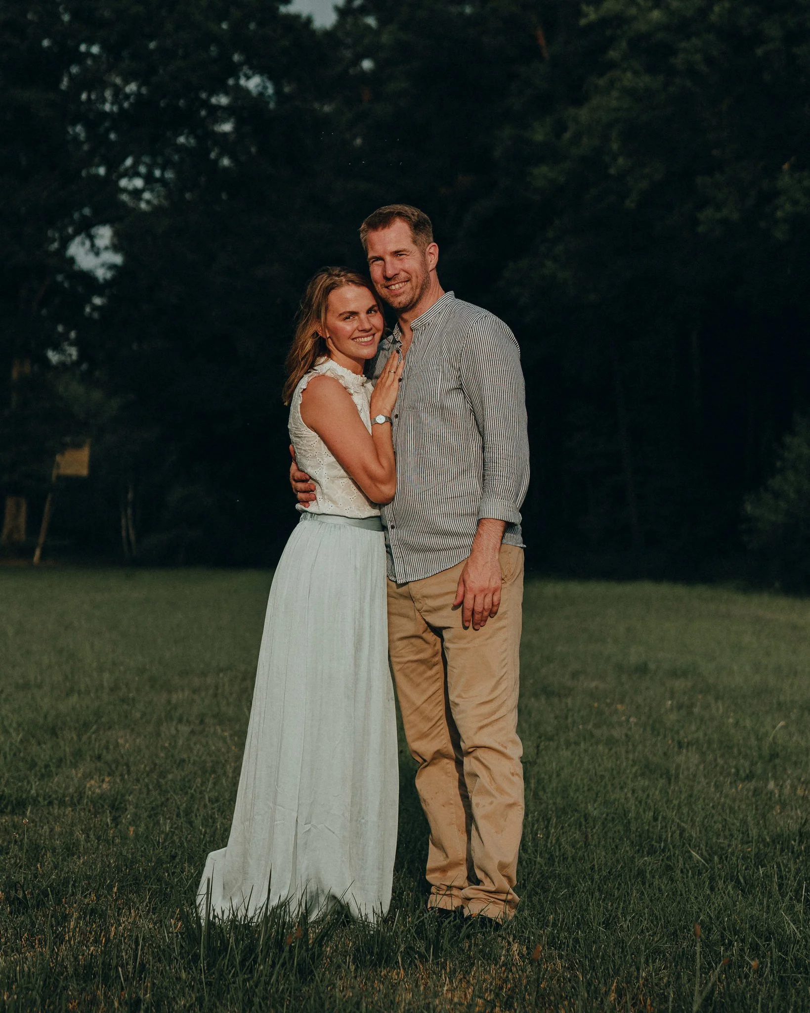 Ein lächelndes Paar steht im Freien auf einer Wiese, umgeben von Bäumen, bei Sonnenuntergang.