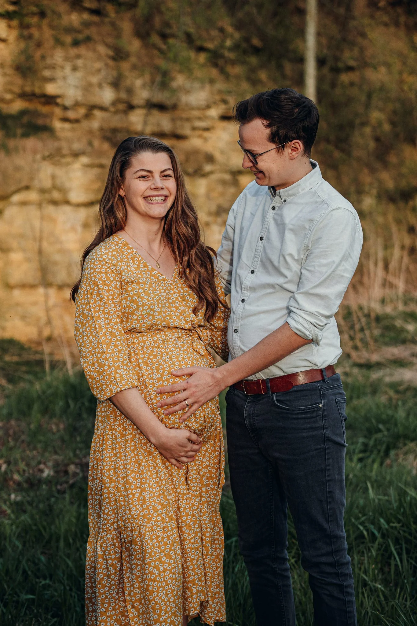 Ein glückliches Pärchen während einer Fotosession im Freien, das eine schwangere Frau zärtlich berührt, umgeben von Natur mit Felsen und Gras, bei Sonnenuntergang.