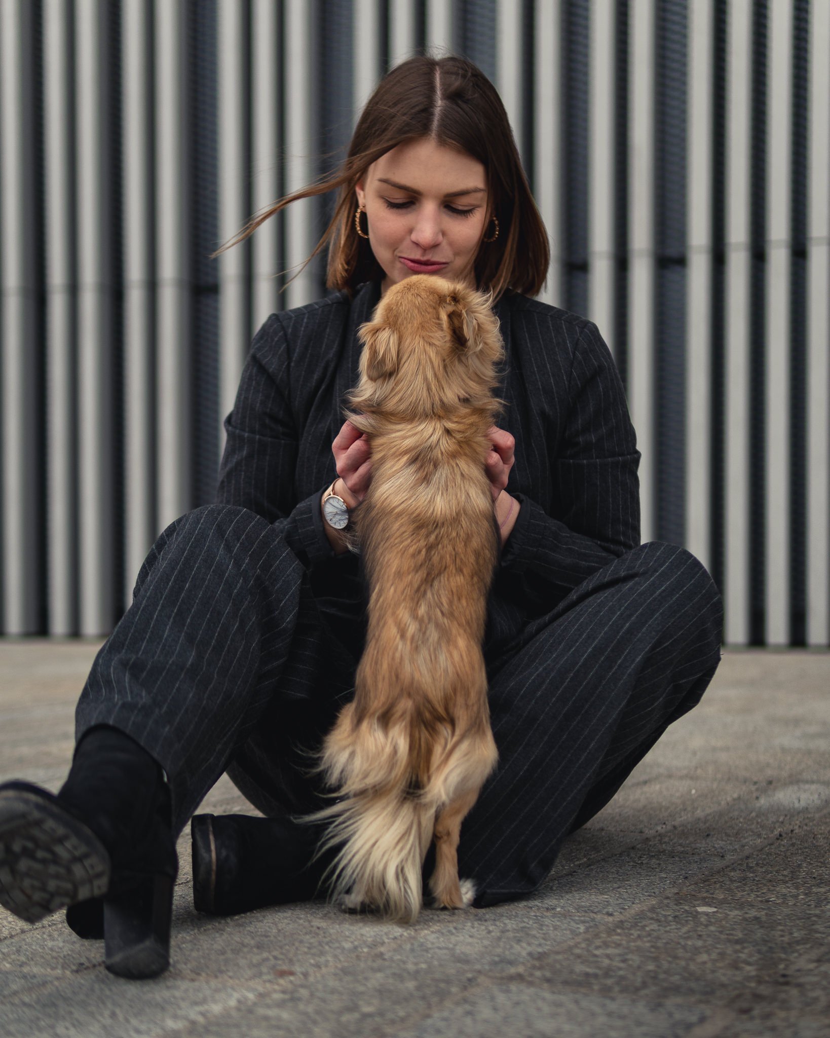 Eine junge Frau in schwarzer Anzugsjacke sitzt auf dem Boden und hält einen kleinen braunen Hund. Sie schauen sich an, vor einem Metallzaun.