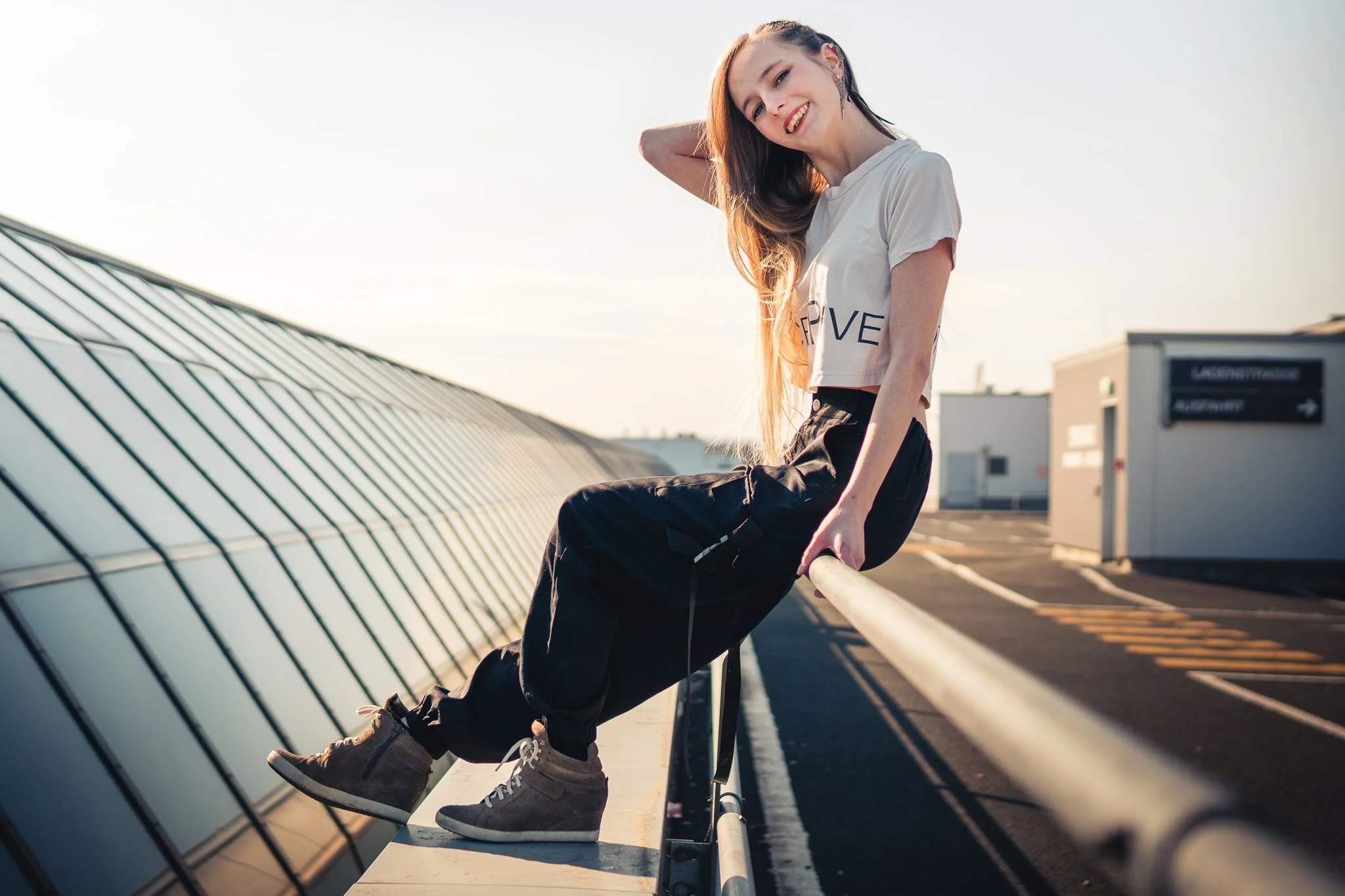 Junge Frau sitzt auf einer Absperrung auf einem Dach bei Sonnenuntergang, lächelnd, in sportlicher Kleidung.