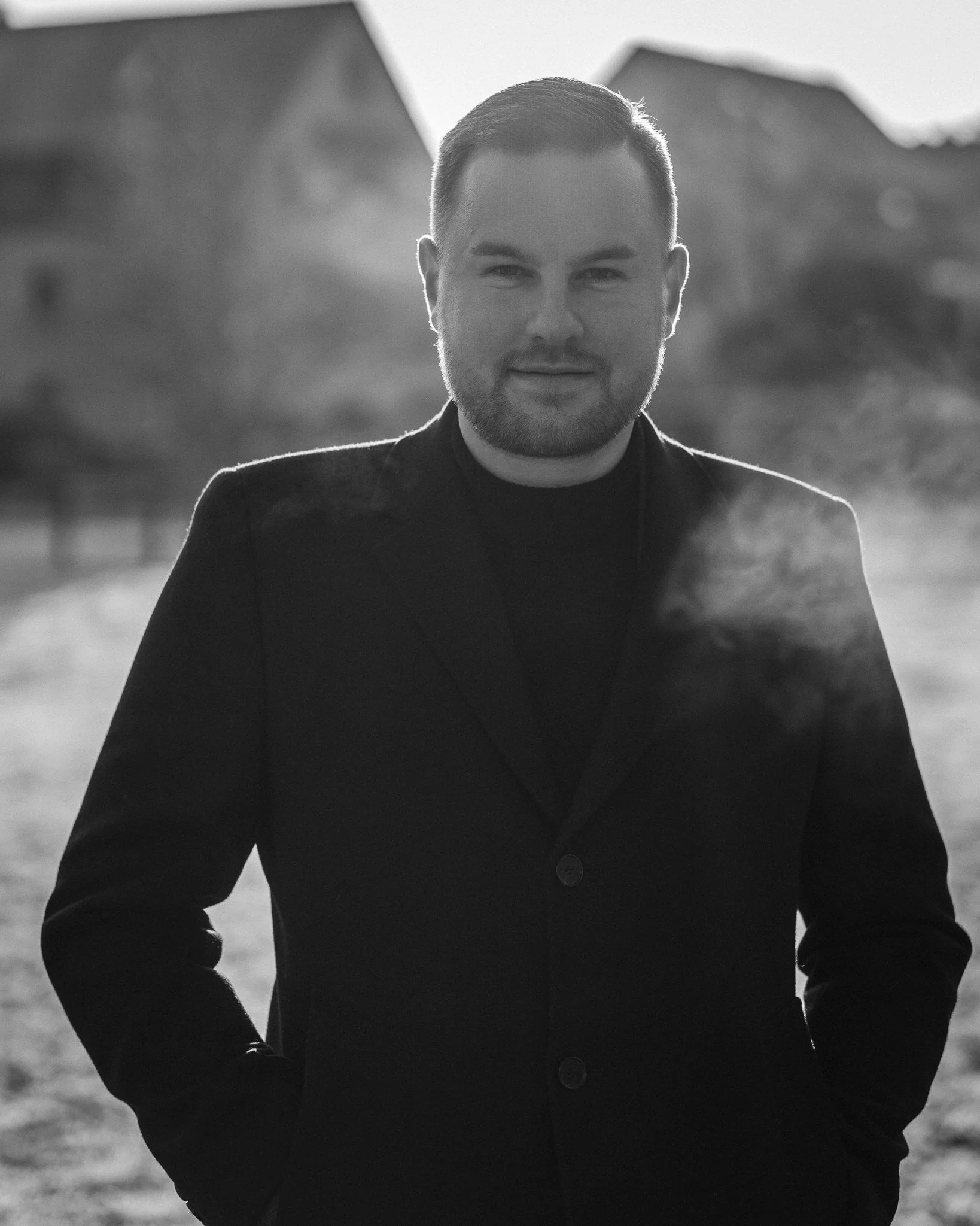 Schwarz-weiß-Fotografie eines Mannes mit Bart, der im Freien steht, trägt eine dunkle Jacke, während die Sonne im Hintergrund untergeht oder aufgeht.