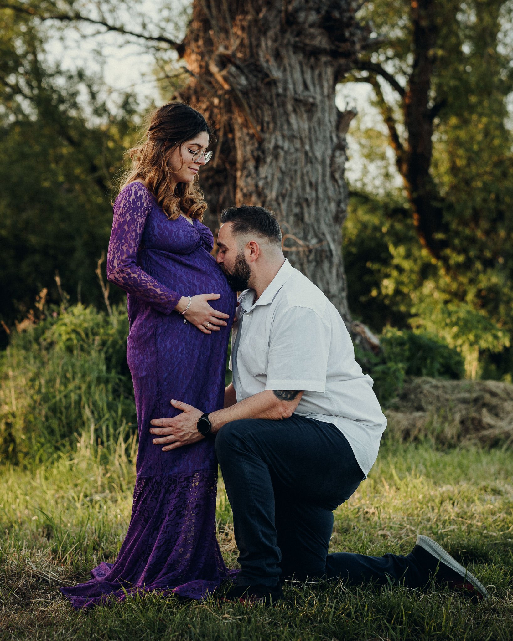 Ein Mann kniet vor einer schwangeren Frau, die in einem lila Kleid steht, und küsst ihren Bauch, vor einem großen Baum im Freien bei Sonnenuntergang.
