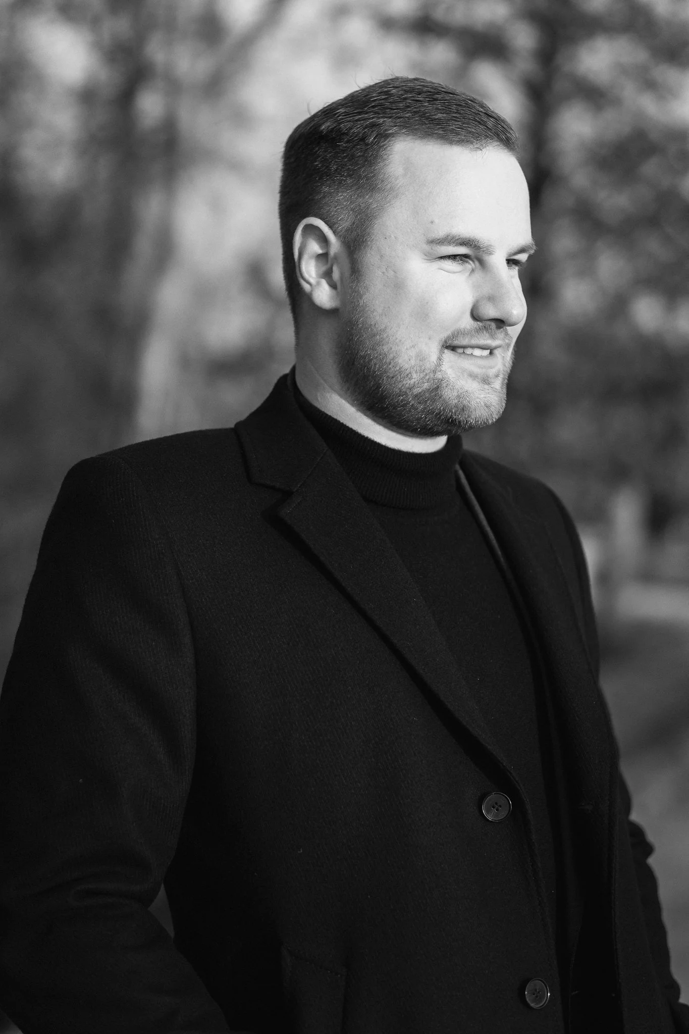 Schwarz-weiß-Fotografie eines Mannes in einem schwarzen Mantel mit kurzer Frisur und Bart, im Freien, lächelnd und in die Ferne blickend.