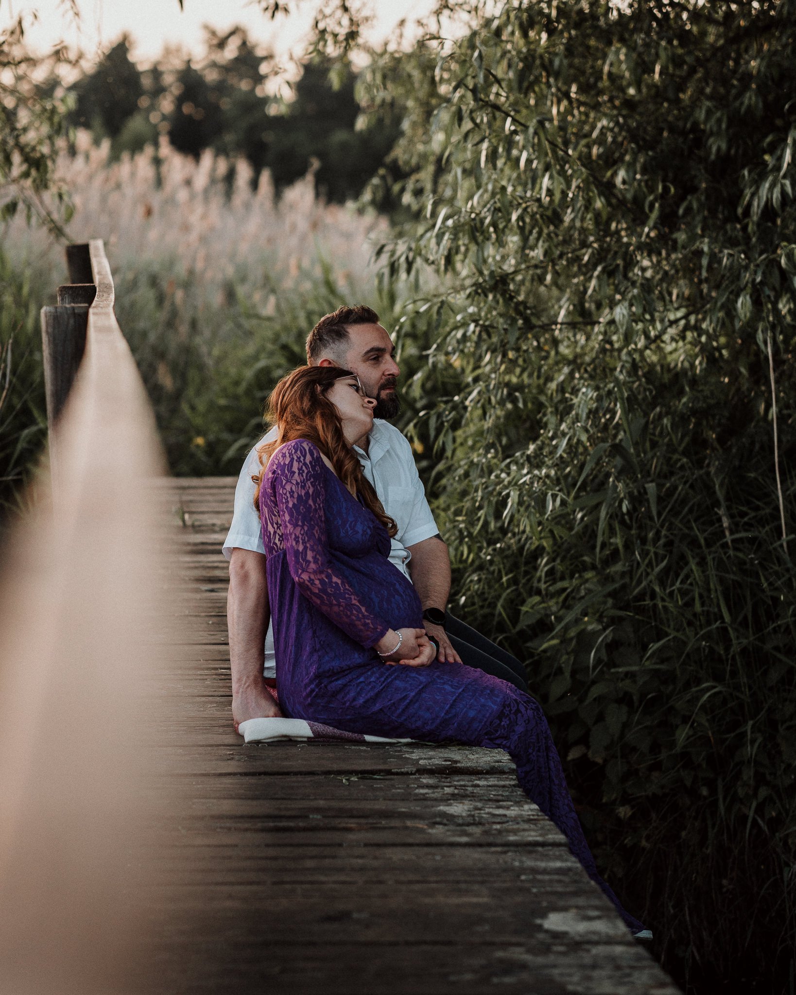 Ein Paar sitzt an einem Steg bei Sonnenuntergang, sie kuschelt sich an ihn, umgeben von Natur und Büschen.