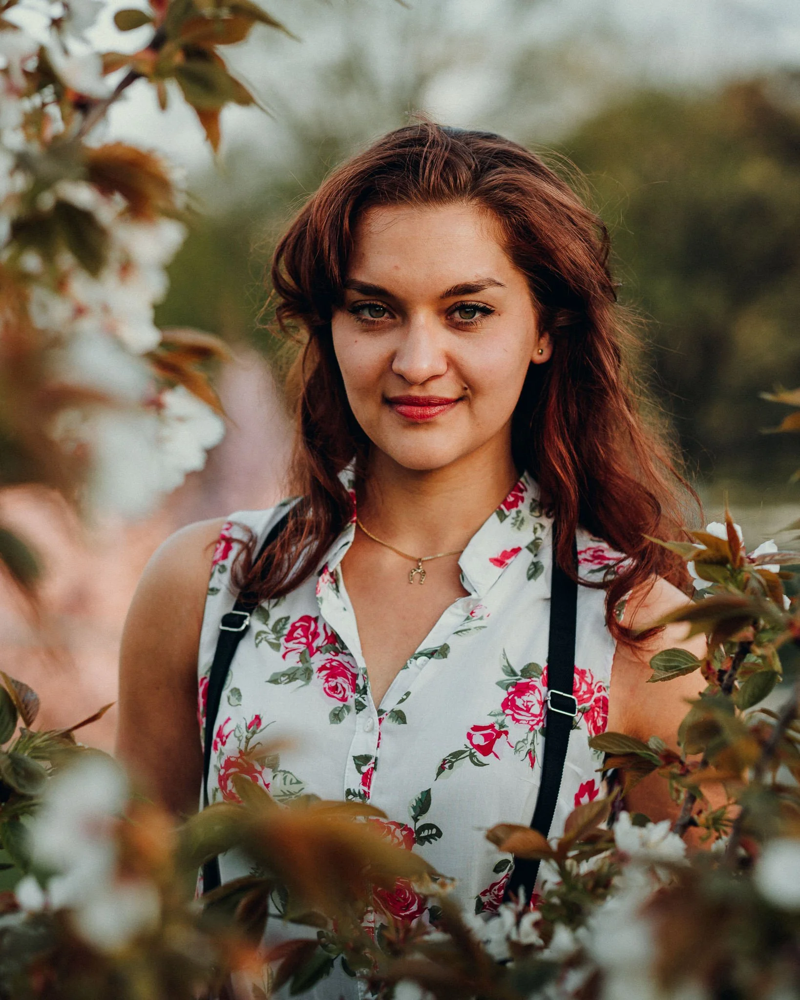 Junge Frau mit roten Haaren in einem blumigen Top, umgeben von Apfelblüten im Freien.