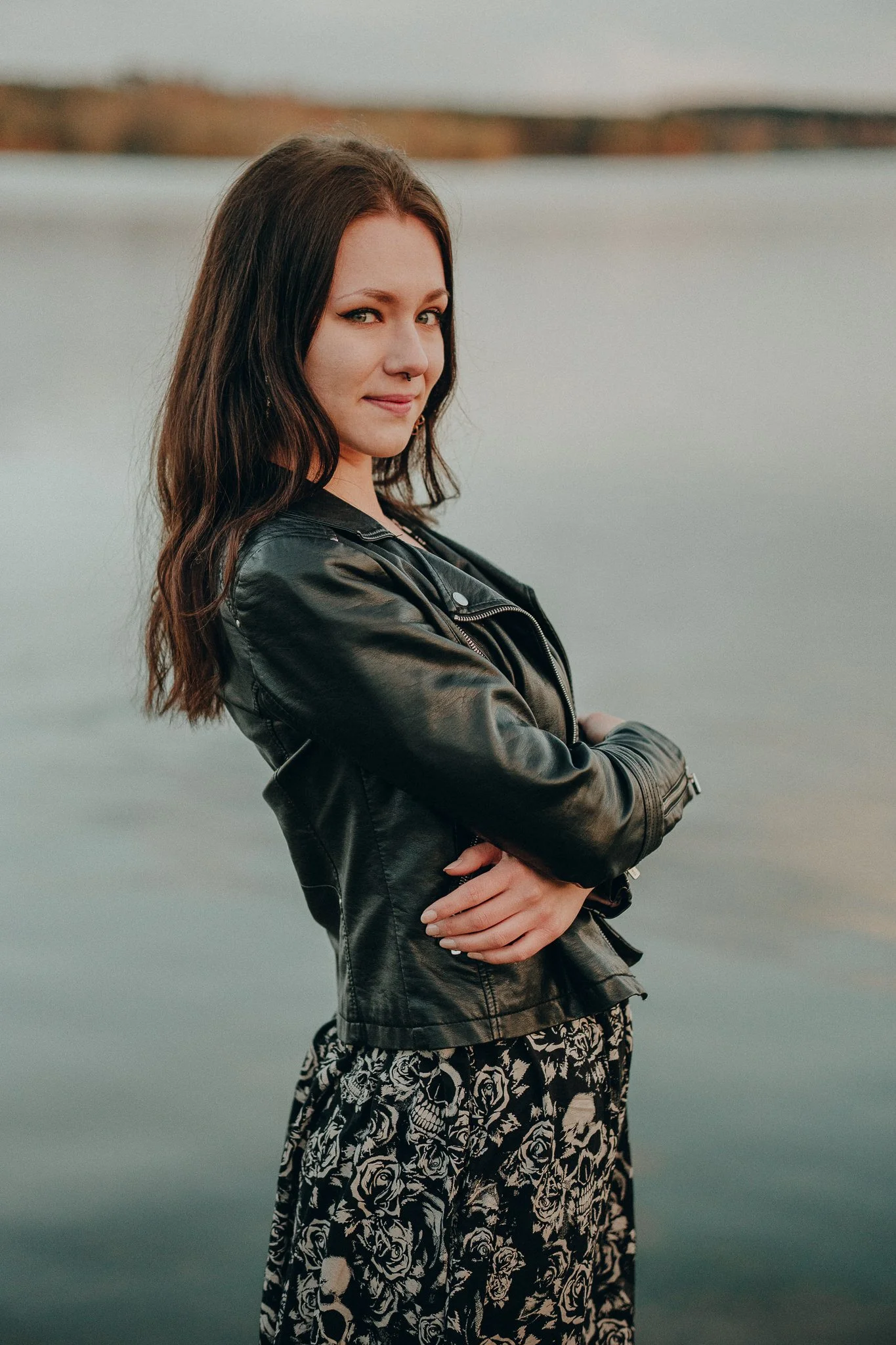 Junge Frau mit dunklen, gewellten Haaren, schwarzer Lederjacke und floralen Rock steht am Wasser und blickt zur Seite.