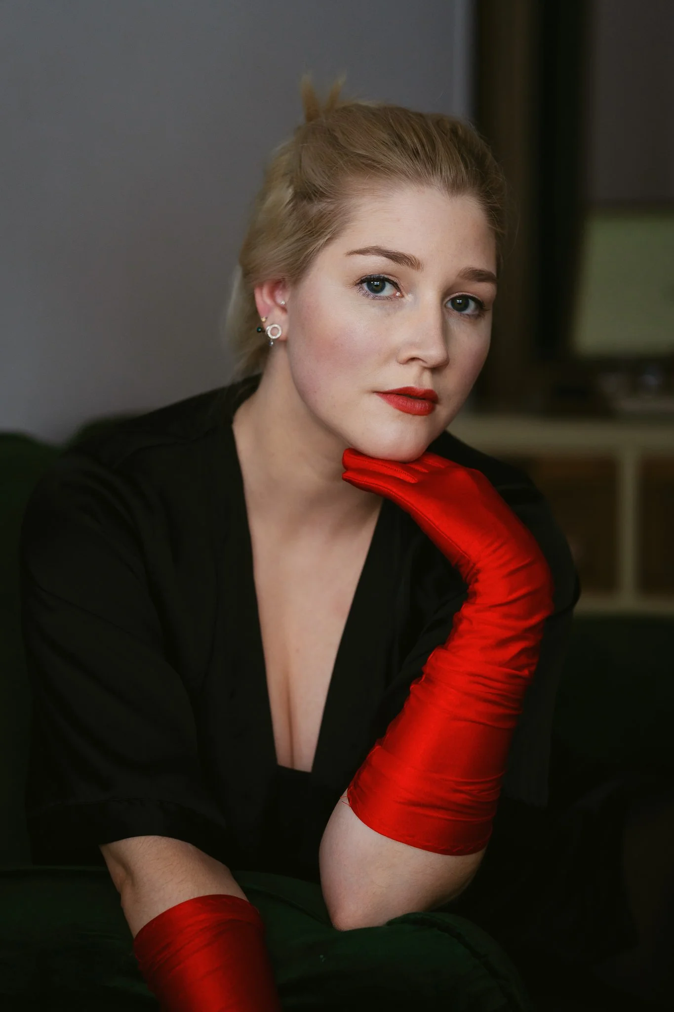 Junge Frau mit blonden Haaren, rotem Lippenstift und roten Handschuhen, die den Kopf mit der Hand stützt, sitzt auf einem Sofa in einem Raum mit dunklem Hintergrund.