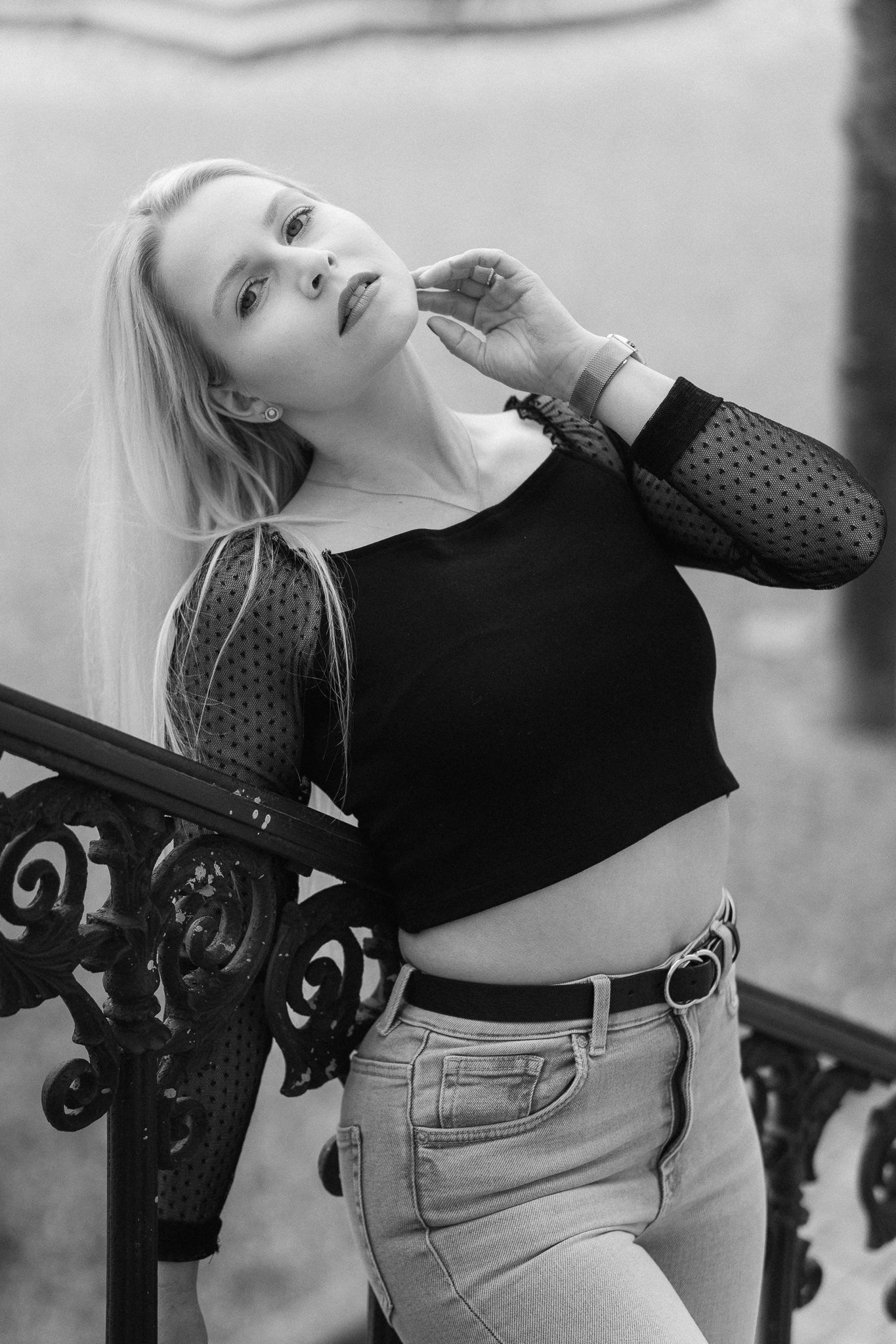 Schwarz-Weiß-Fotografie einer jungen Frau mit langen Haaren, die zu einer Seite blickt, mit einer Hand am Gesicht und einer modernen schwarzen Bluse mit transparenter, gepunkteter Manschette, Jeans und einem Gürtel. Im Hintergrund eine dekorative Met