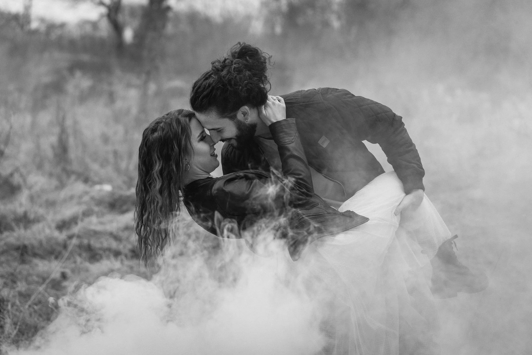 Ein Paar, das sich in einer grauen, rauchigen Umgebung im Freien liebevoll umarmt und küsst, getragen von Gefühlen der Nähe und Intimität, in Schwarz-Weiß-Fotografie.