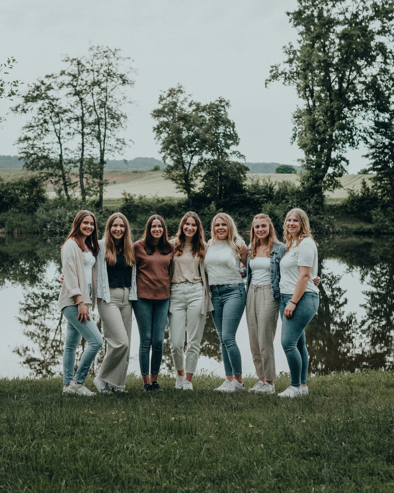 Eine Gruppe von sieben jungen Frauen steht dicht beieinander am Ufer eines Sees in der Natur, umgeben von Bäumen und grünen Hügeln im Hintergrund.