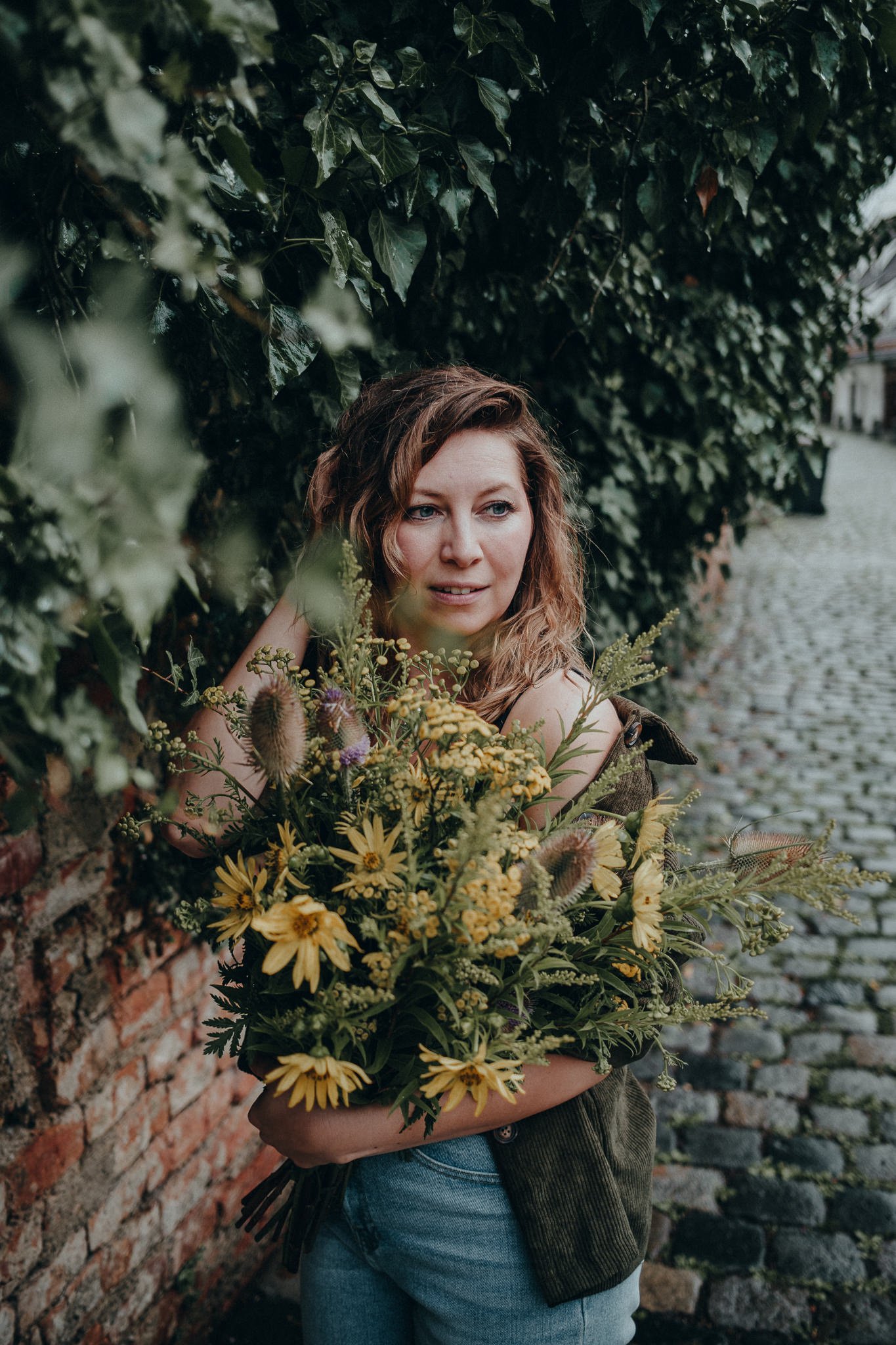 Eine Frau mit braunen, lockigen Haaren hält einen großen Blumenstrauß mit gelben, lila und grünen Blumen, steht neben einer alten Ziegelmauer und grünen Büschen im Hintergrund auf einer gepflasterten Straße.