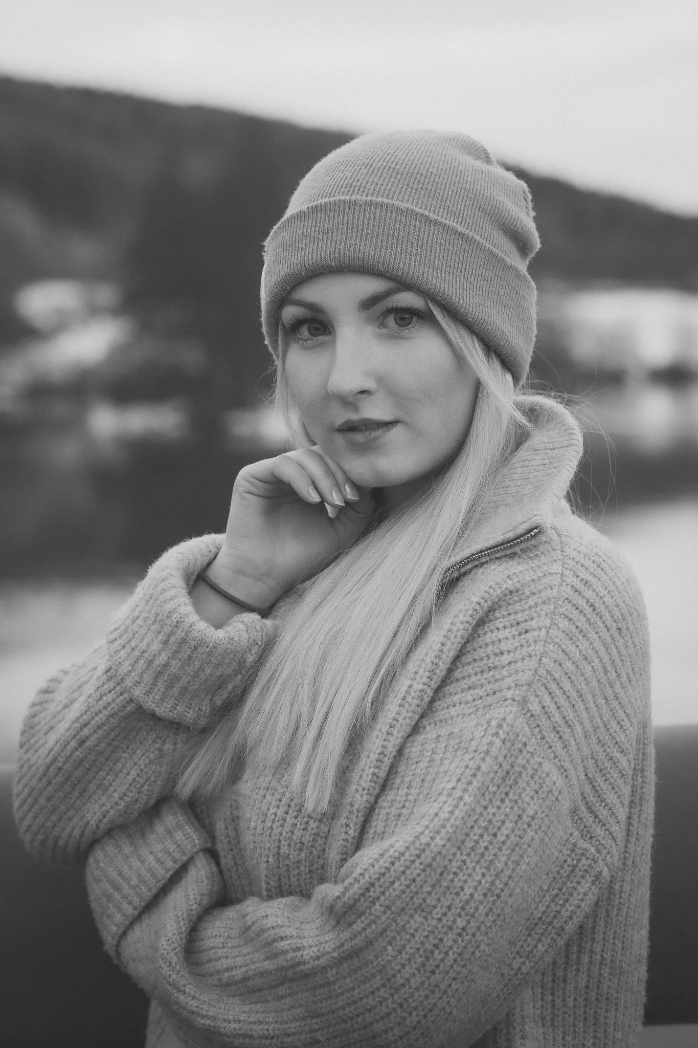 Schwarz-Weiß-Fotografie eines jungen Mannes mit blondem Haar, der einen Beanie trägt und in die Kamera blickt, mit einem See und Bergen im Hintergrund.