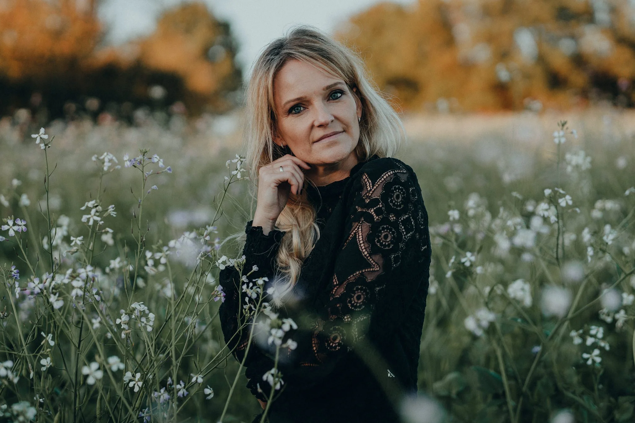 Eine blonde Frau in einem schwarzen Spitzenkleid steht in einem Feld mit weißen und violetten Blumen bei Sonnenuntergang.