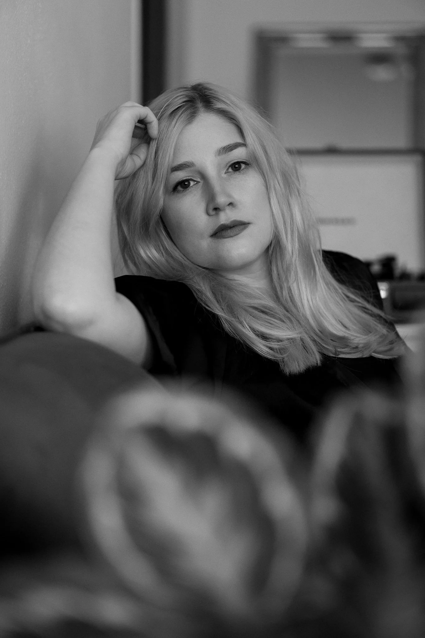 Schwarzweißfoto einer jungen Frau mit blonden, lockigen Haaren, die auf einem Sofa sitzt und in die Kamera schaut, mit einem nachdenklichen Ausdruck.