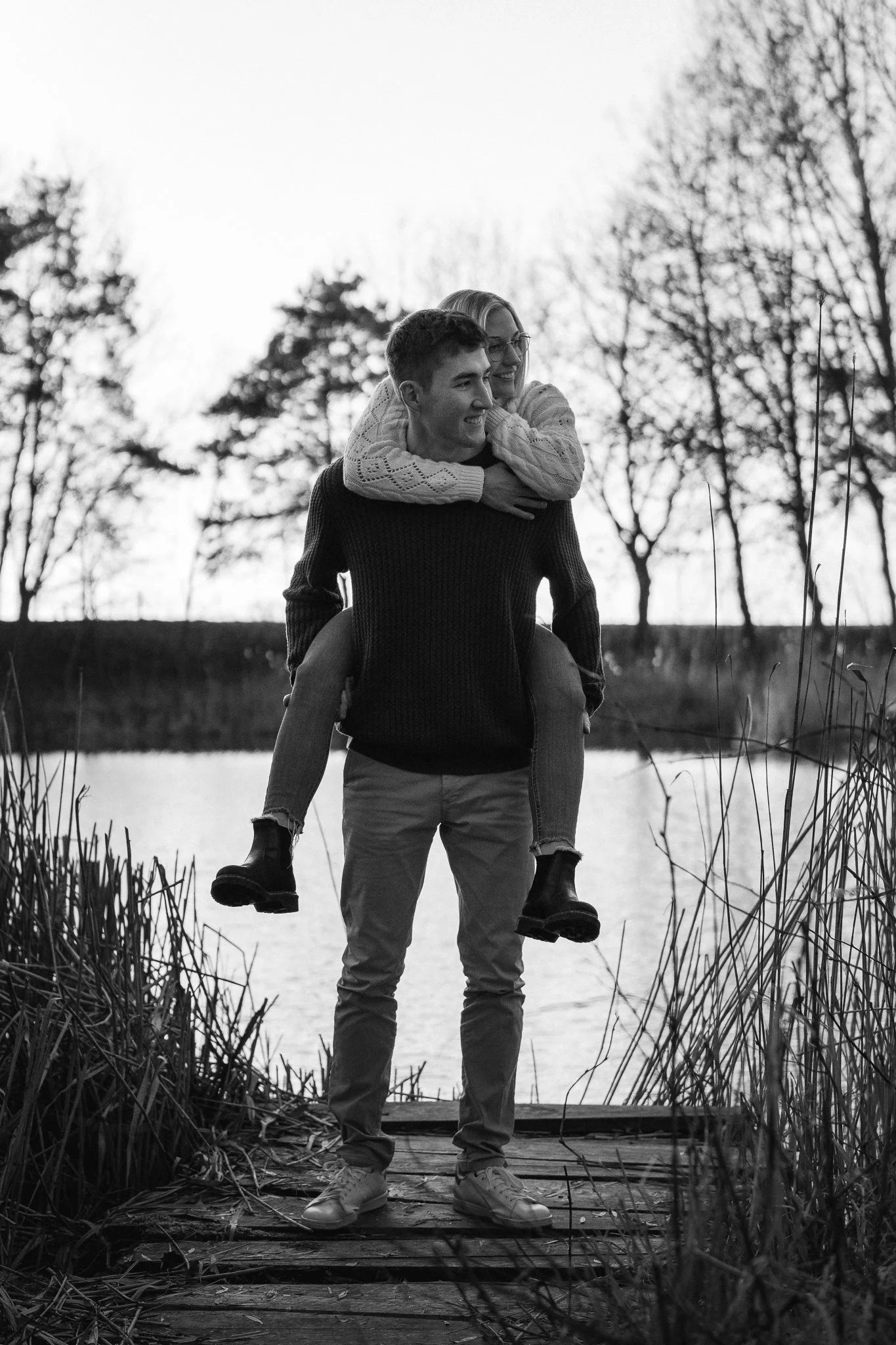 Ein Paar beim Spaziergang am See, der Mann trägt eine dunkle Jacke und helles Hosen, während die Frau auf seinem Rücken sitzt, lächelnd und um seinen Hals gewickelt. Es ist schwarz-weiß.