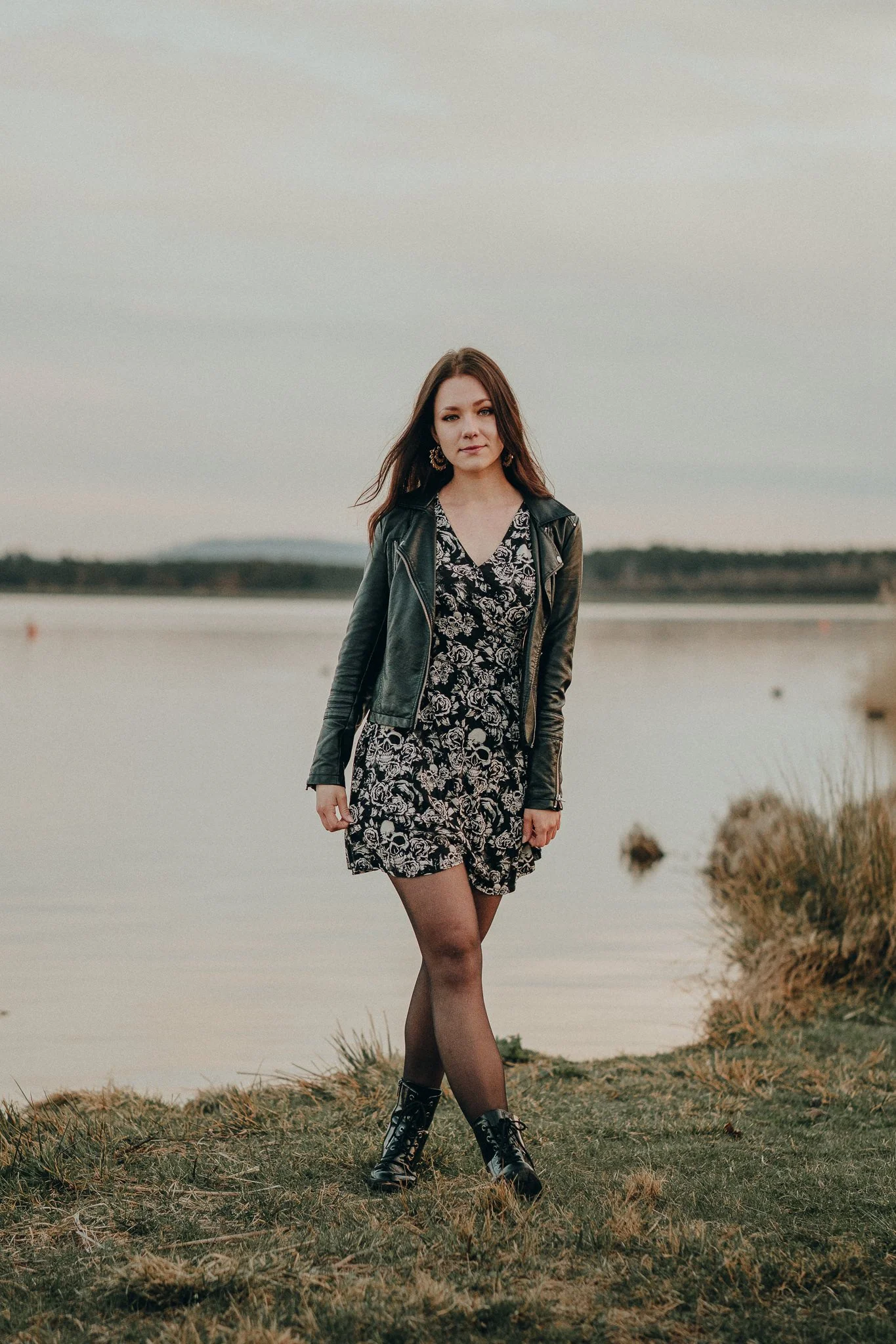 Eine junge Frau steht am Ufer eines Sees bei Sonnenuntergang, trägt ein schwarzes Lederjacke, ein schwarzes Kleid mit weißen Totenköpfen, schwarze Strümpfe und schwarze Stiefel.