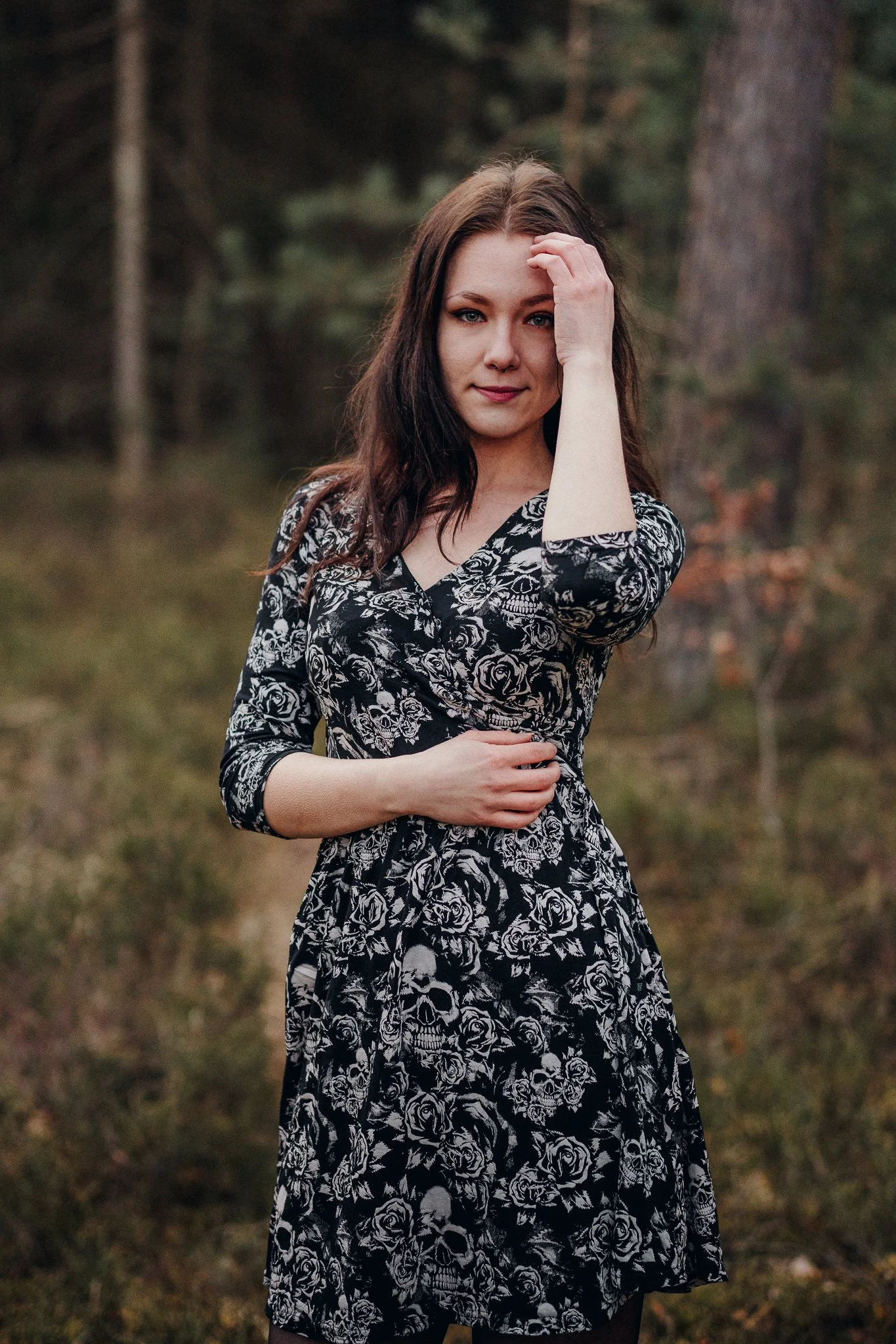Junge Frau mit schwarzem Kleid mit Schädel- und Rosenmuster steht in einem bewaldeten Gebiet, ihre linke Hand berührt ihr Haar, während sie in die Kamera schaut.