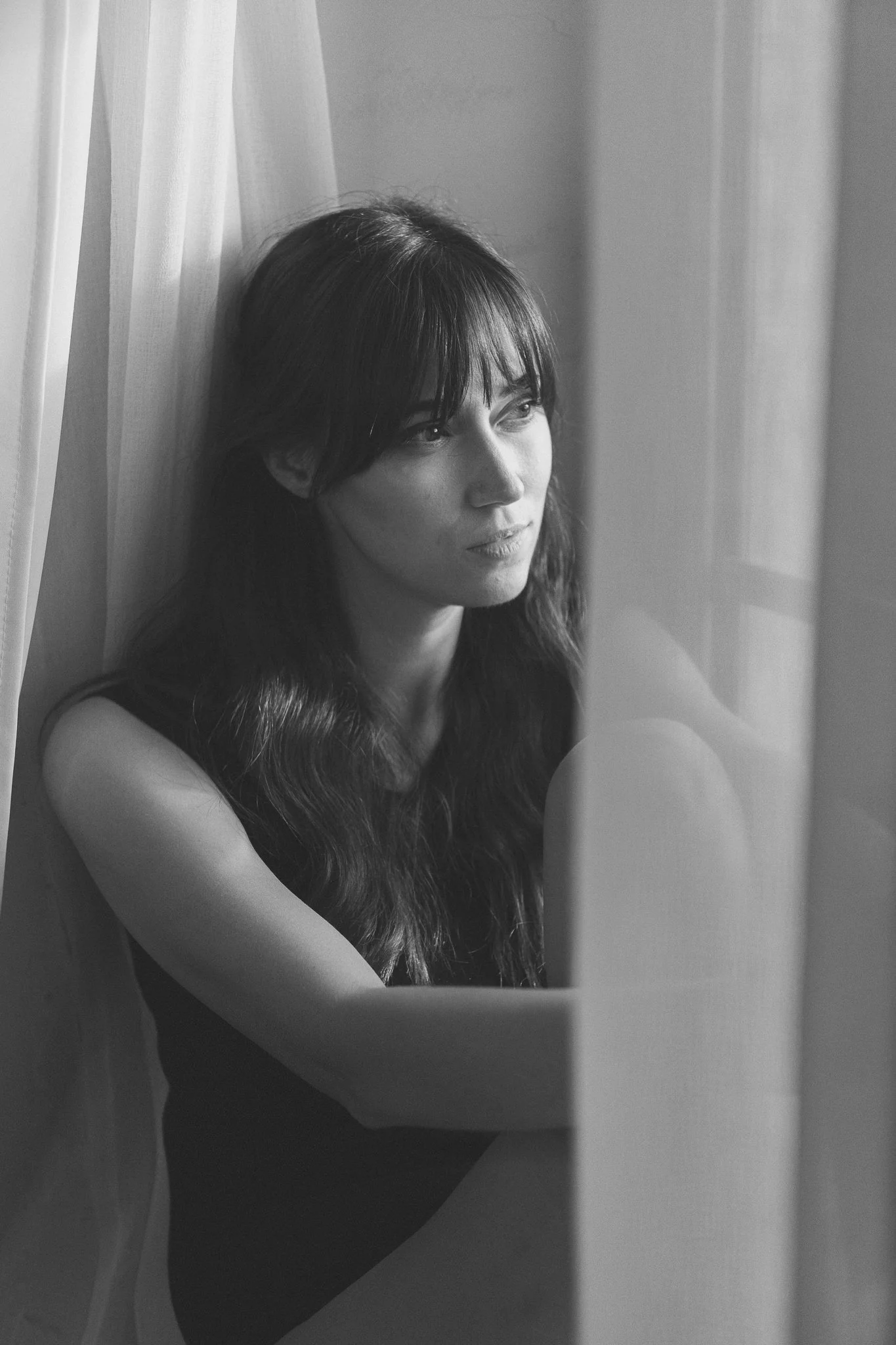 Schwarz-weiß-Foto einer Frau mit langen dunklen Haaren, die neben einem Fenster sitzt und nach draußen blickt.
