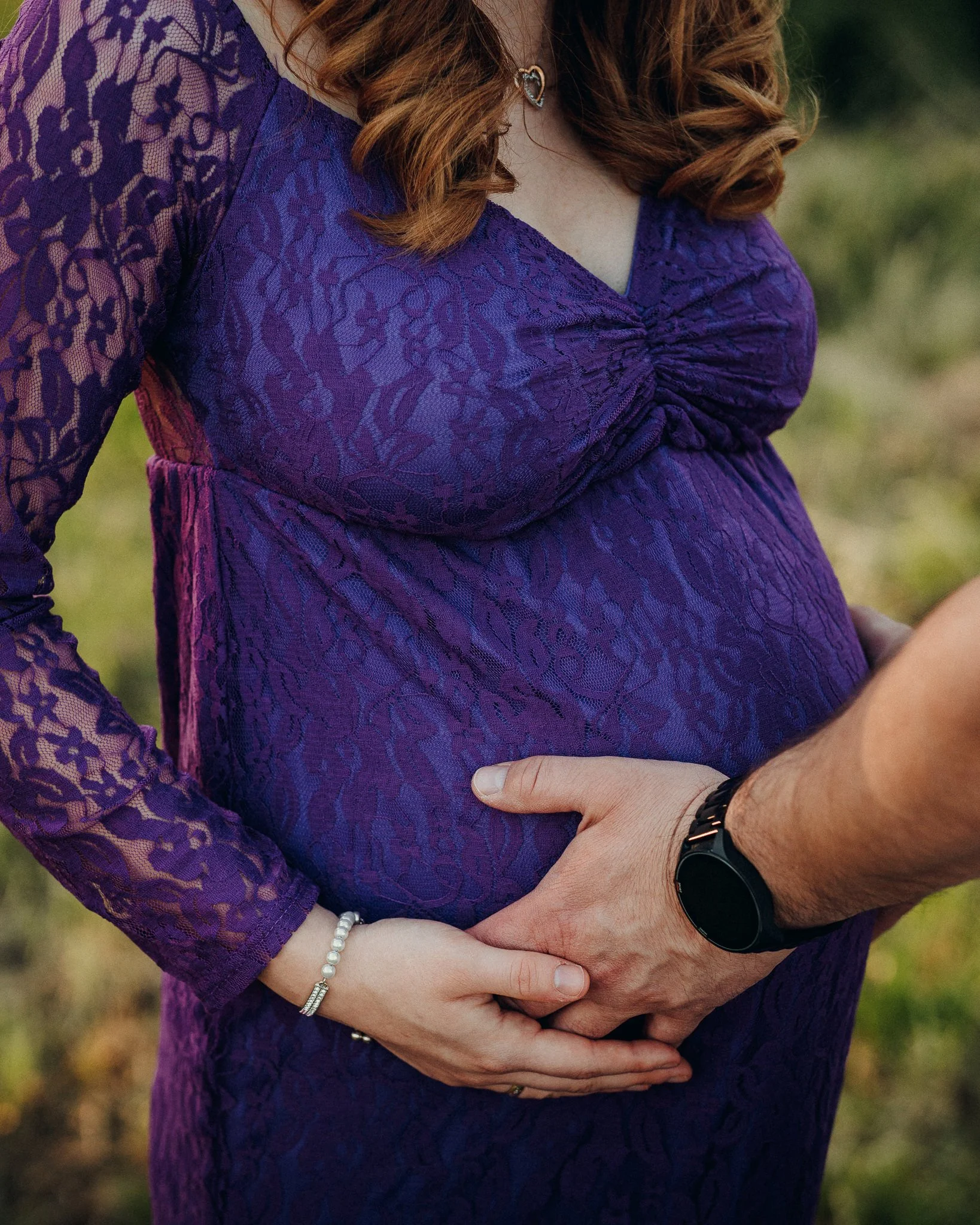 Nahaufnahme eines schwangeren Frauenbauchs in einem lila Spitzenkleid, gehalten von zwei Händen, im Freien, mit sichtbaren Schmuckstücken und einem schwarzen Armband.
