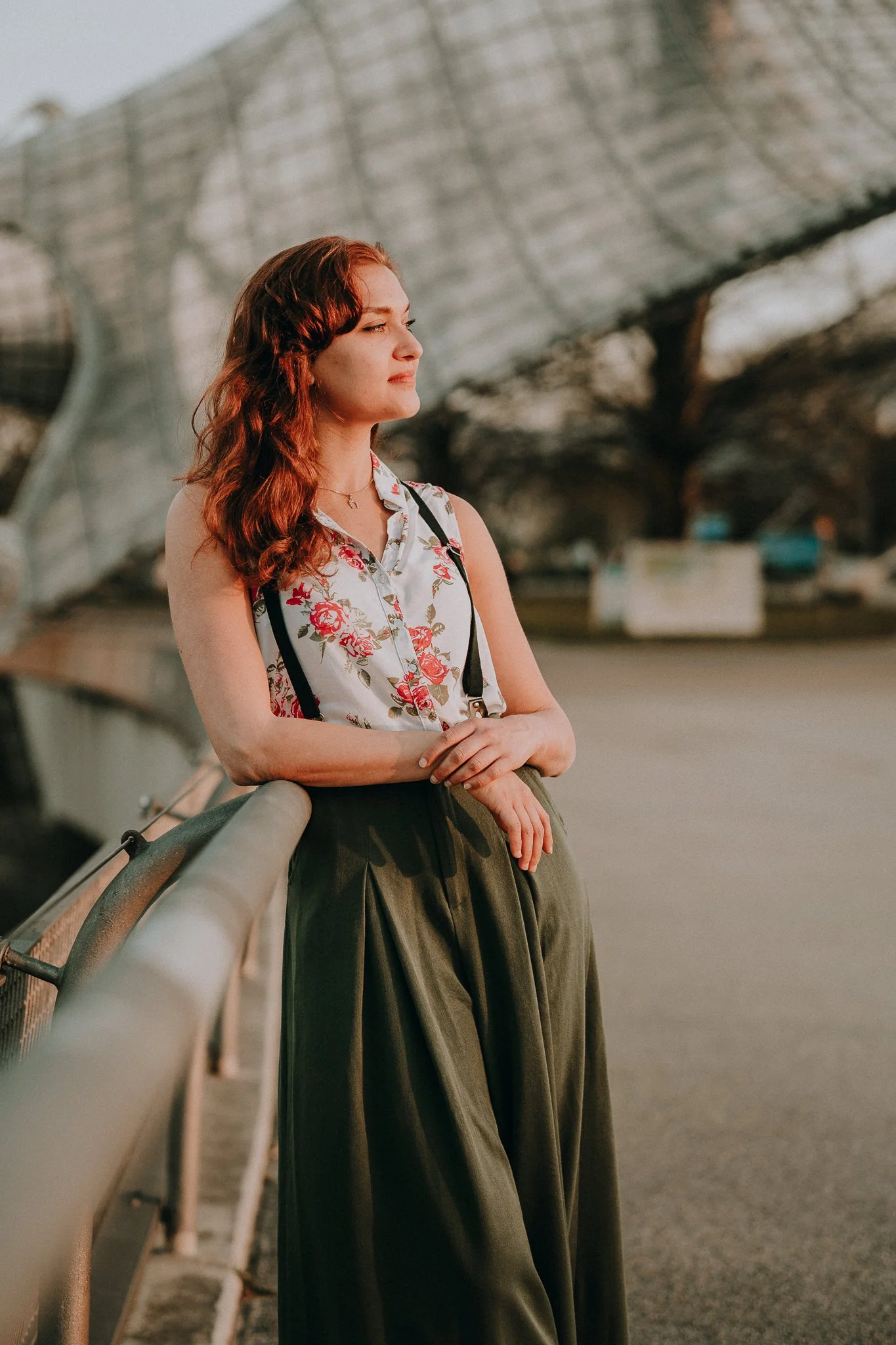 Junge Frau mit roten, gewellten Haaren steht an einem Geländer und blickt nach links, im Hintergrund eine moderne Brücke bei Sonnenuntergang.