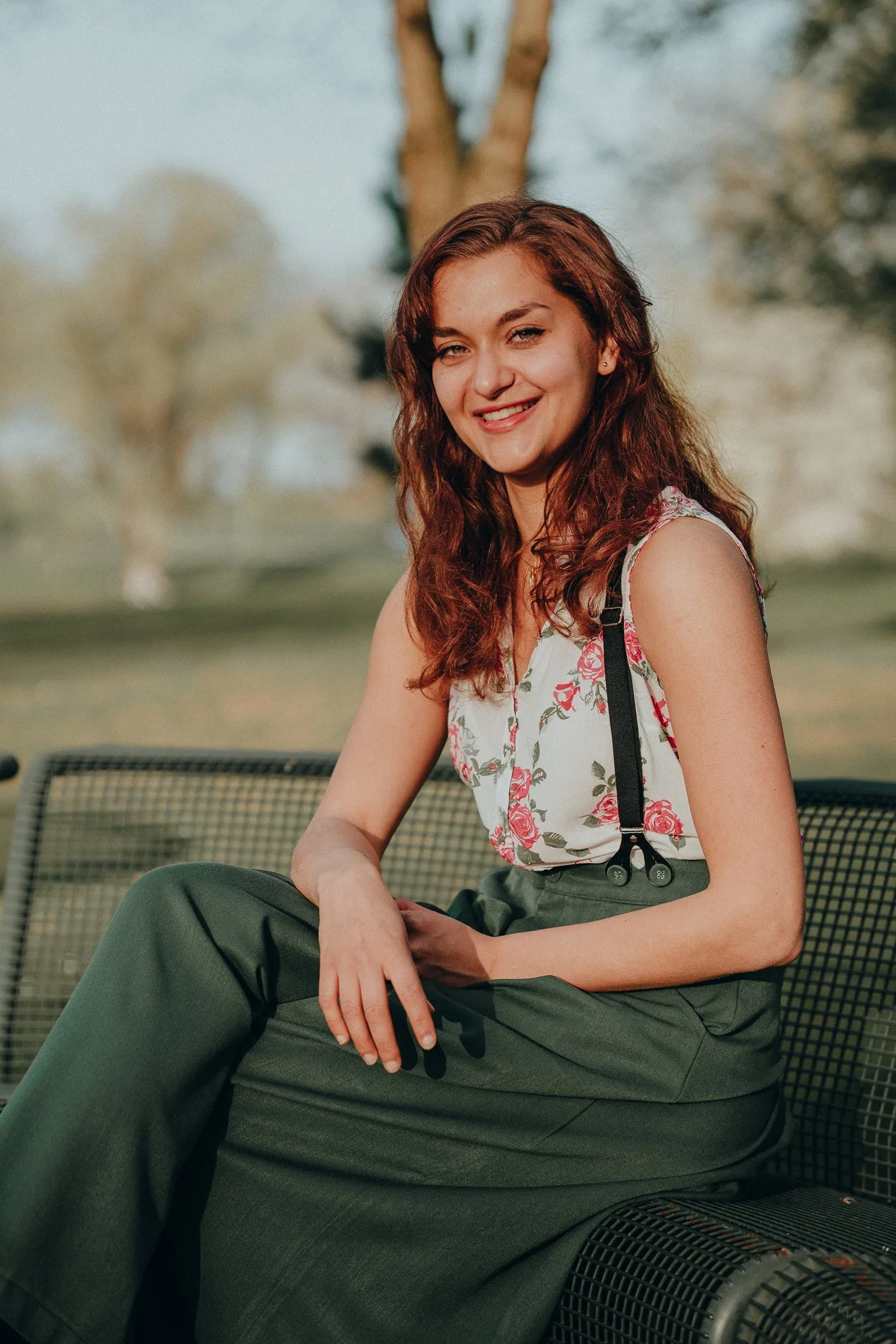 Junge Frau mit roten Haaren sitzt auf einer Parkbank im Freien und lächelt in die Kamera, im Hintergrund Bäume.