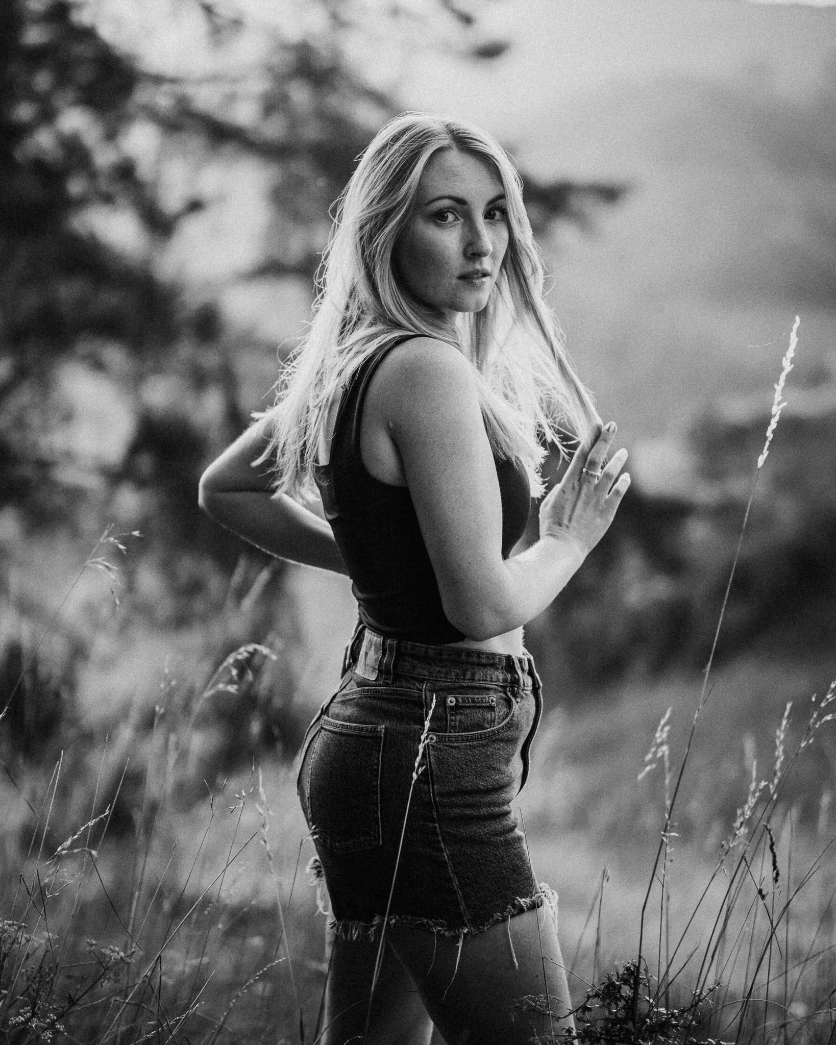 Schwarz-weiß-Foto eines jungen Mädchens, das in einem Feld steht, mit einer Hand auf der Brust, die andere am Rücken, Blick leicht zur Seite gerichtet, mit lockigem, langen Haar.