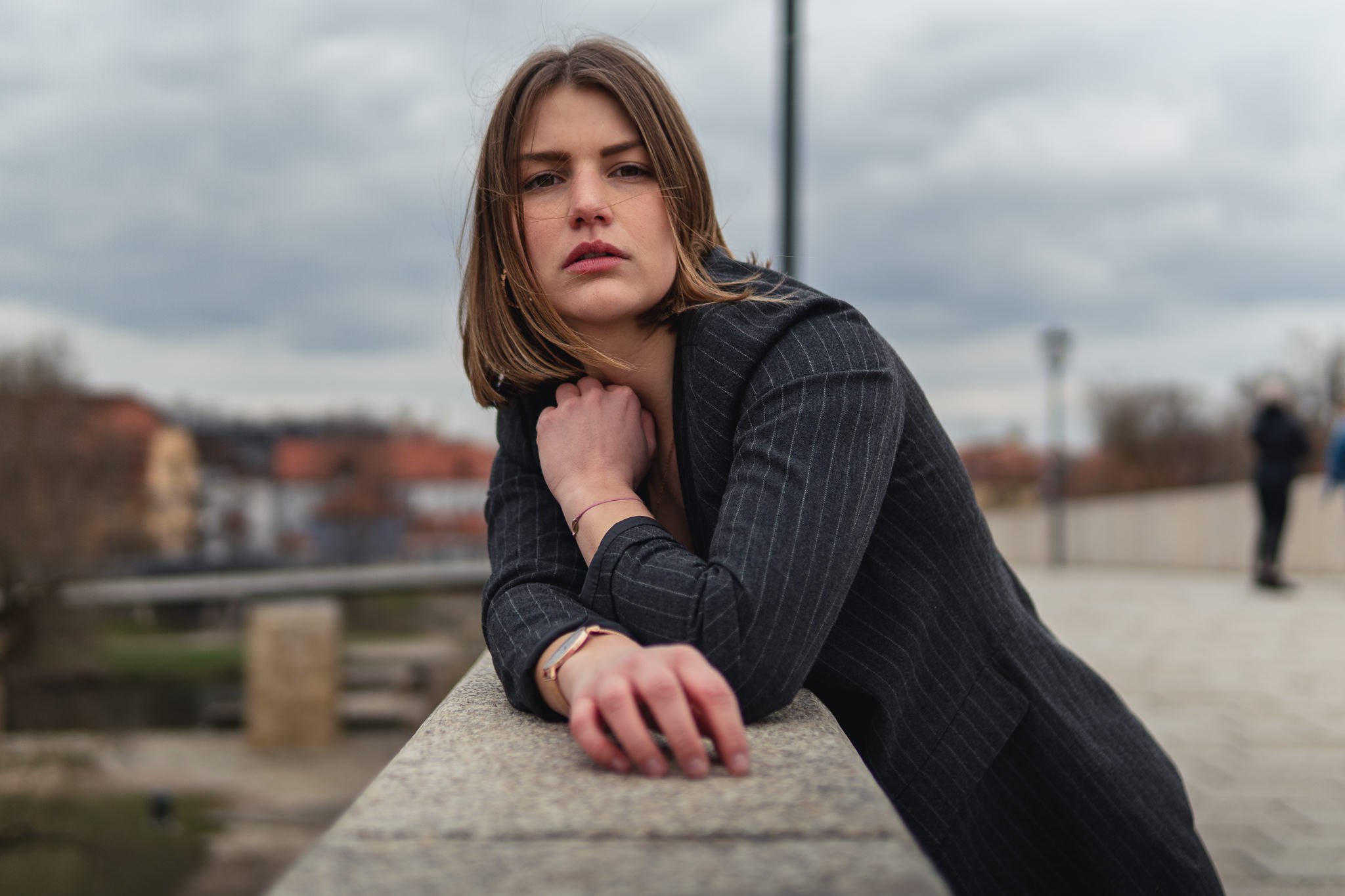 Eine junge Frau mit braunen Haaren und dunklem Blazer lehnt auf einer Brüstung im Freien bei bewölktem Himmel, blickt ernst in die Kamera.