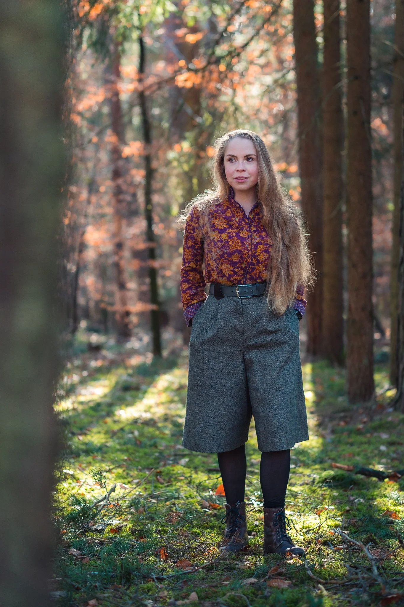 Eine junge Frau mit langen, gewellten Haaren steht in einem Wald im Herbst, trägt eine bunte Bluse, eine weite graue Hose, schwarze Leggings und Stiefel, Blick nach links gerichtet, im Sonnenlicht mit herbstlichen Bäumen im Hintergrund.