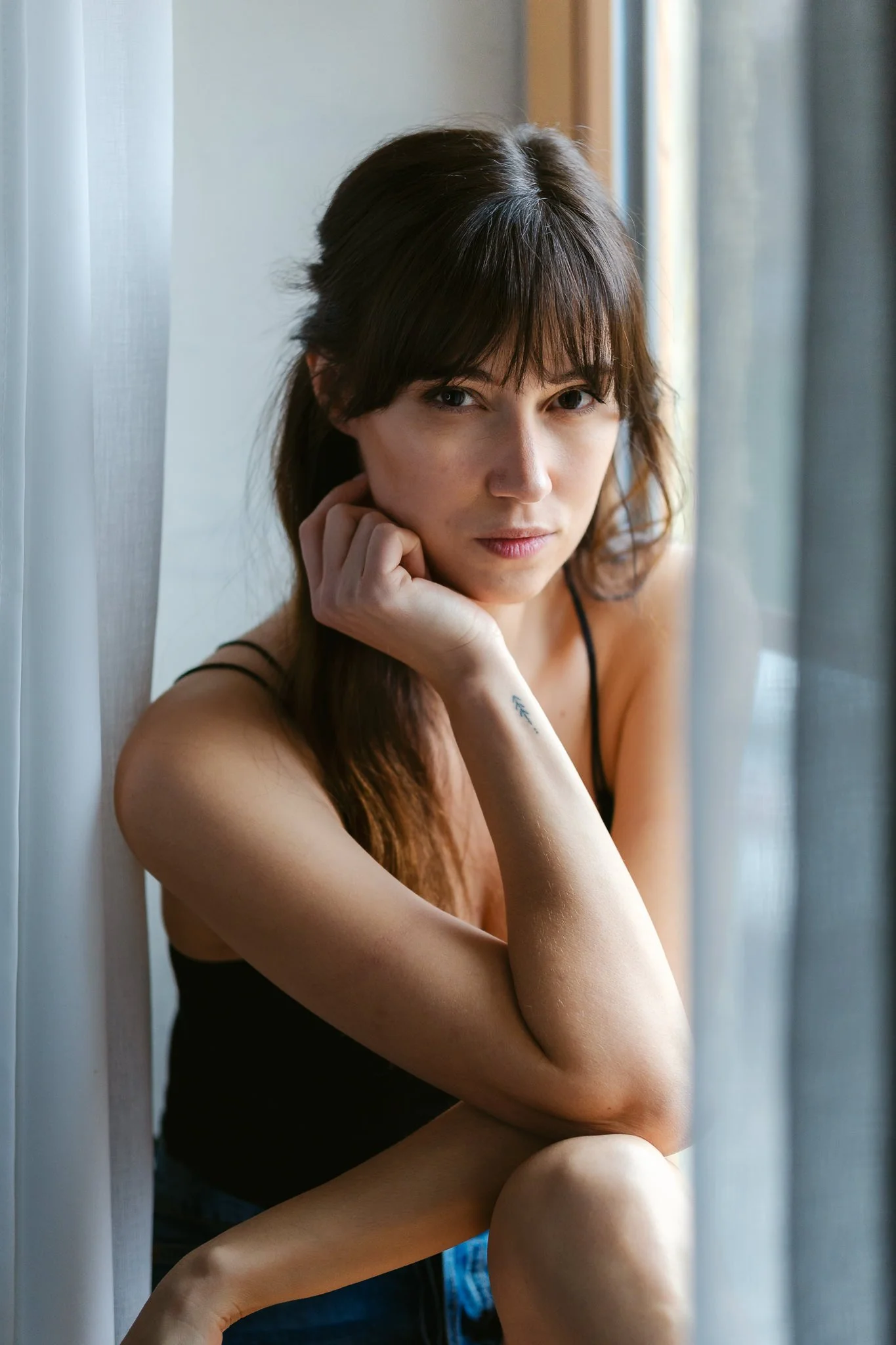 Junge Frau mit braunen Haaren sitzt neben Fenster, trägt schwarzes Top, blickt nach vorne, ruht Hand am Kinn, neben ihr ist ein Vorhang.
