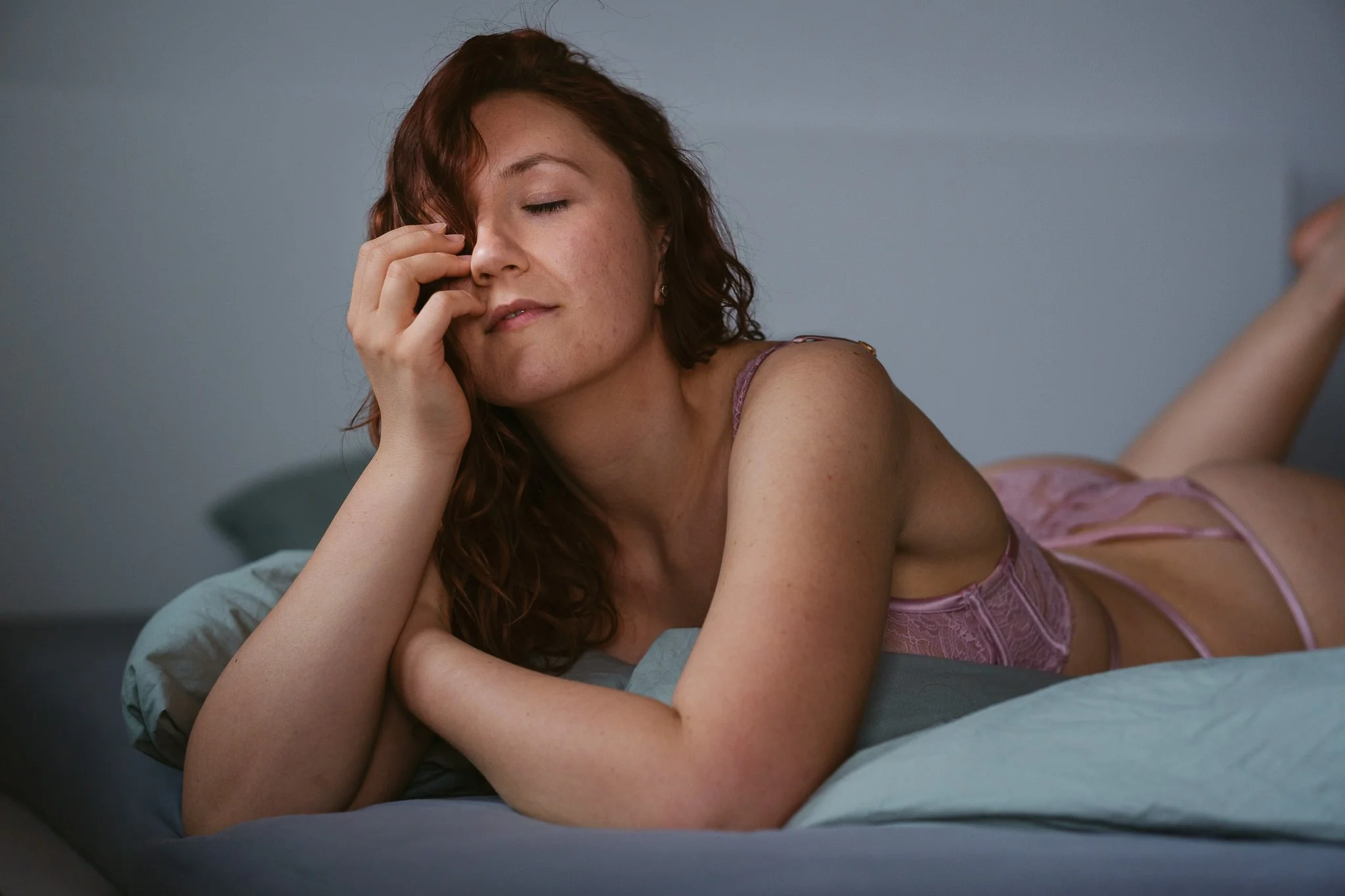 Frau liegt auf einem Bett, legt den Kopf auf den Arm, trägt lila Unterwäsche, hat rot braune, lockige Haare, scheint entspannt und nachdenklich zu sein.