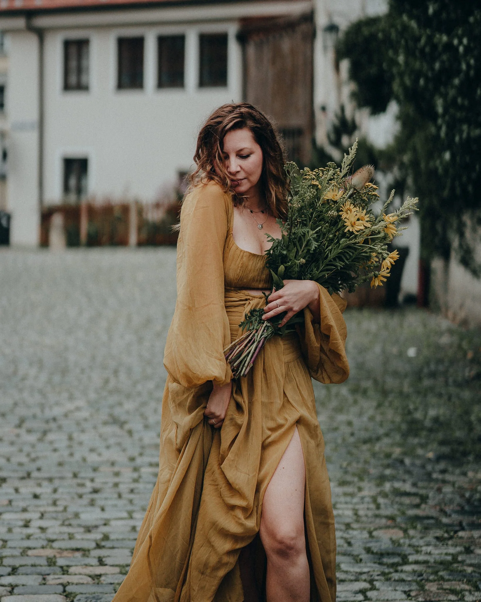 Eine Frau mit braunen, lockigen Haaren trägt ein langes, gelbes Kleid und hält einen Blumenstrauß aus gelben und grünen Pflanzen, während sie auf einer gepflasterten Straße im Freien steht.