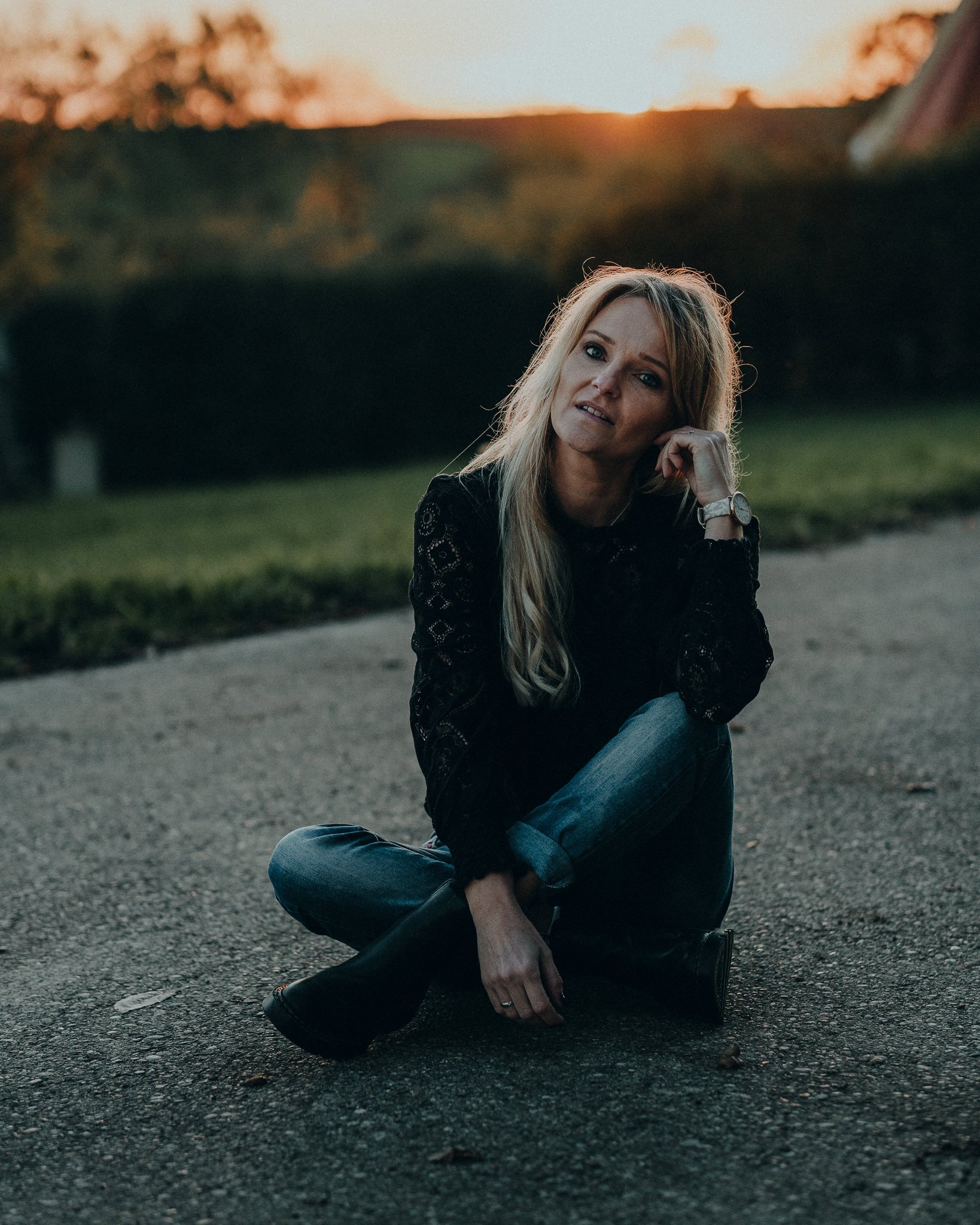 Eine Frau sitzt auf dem Asphalt bei Sonnenuntergang, trägt eine schwarze Spitze-Bluse, Jeans und Stiefel, blickt nach oben mit nachdenklicher Miene.