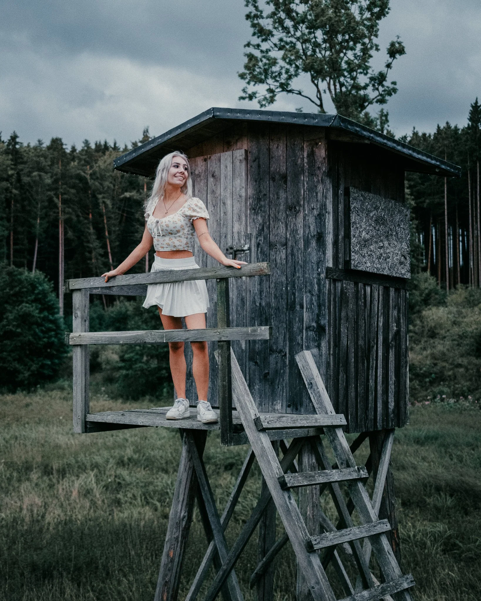 Ein junges Mädchen steht auf einer Holzleiter vor einem hölzernen Vogelhaus im Freien, umgeben von Bäumen und Wäldern