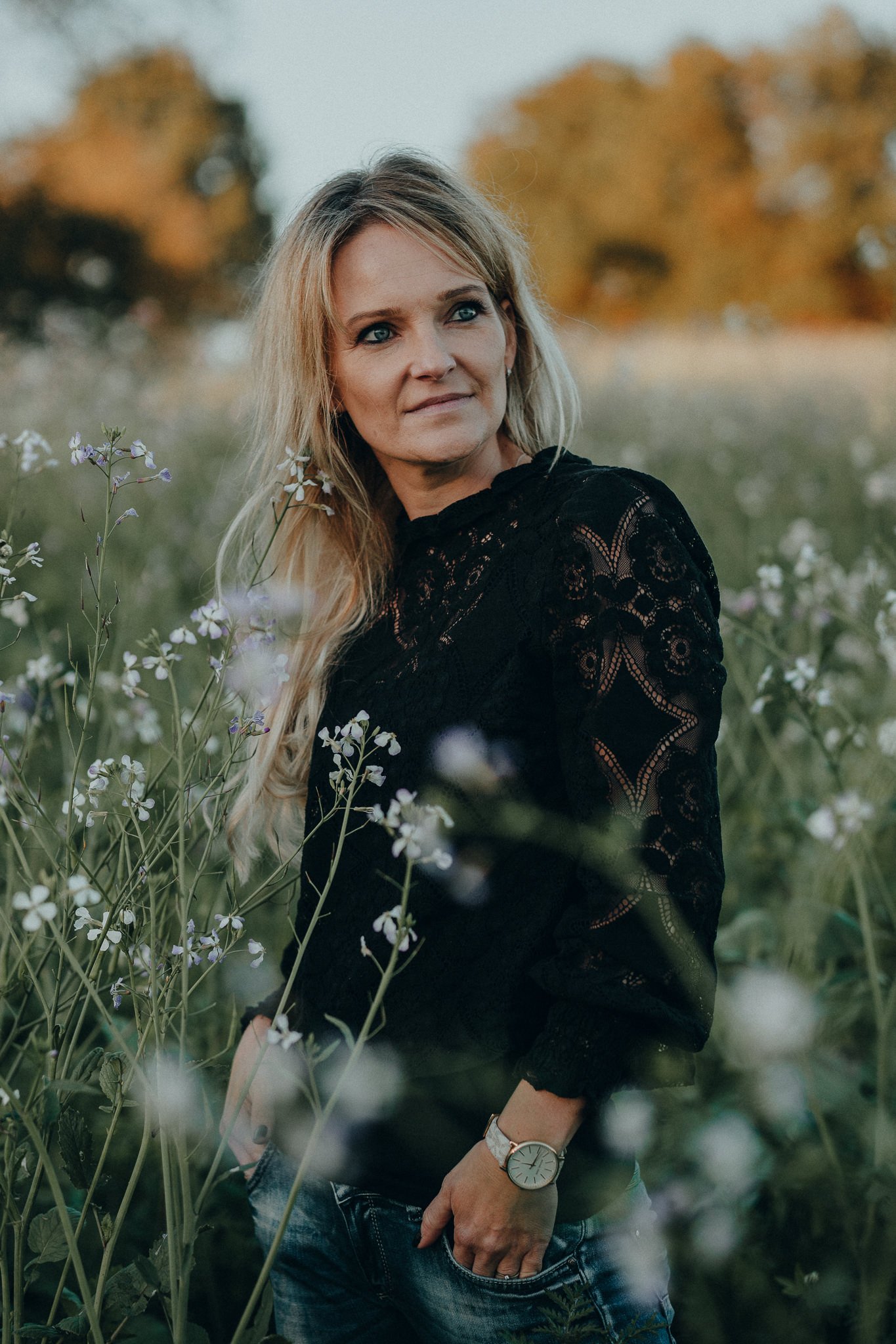 Eine blonde Frau trägt eine schwarze Spitzenbluse und eine Armbanduhr, steht in einem Blumenfeld und blickt seitlich in die Ferne.