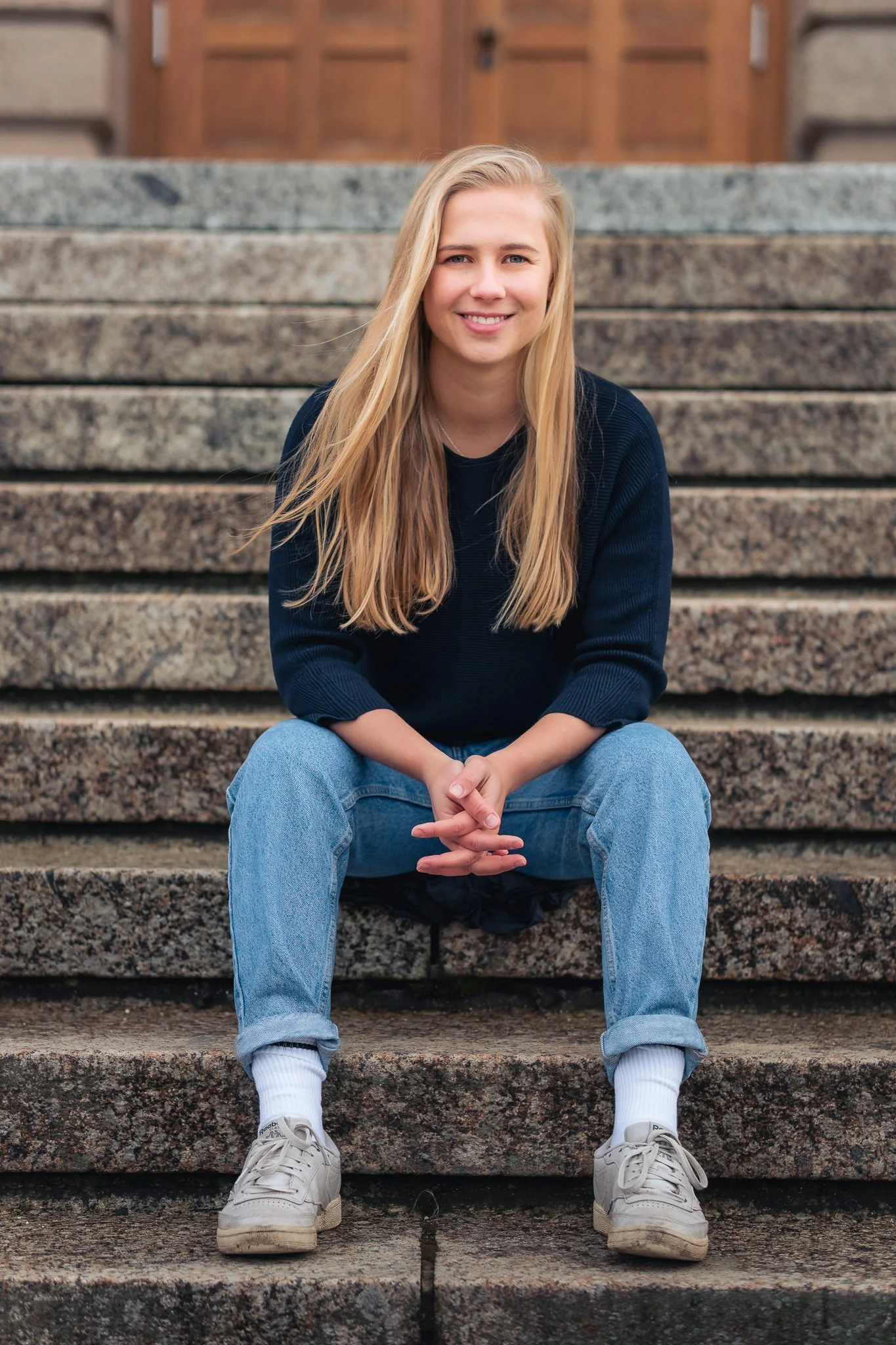 Junge Frau sitzt auf Steintreppe, lächelnd, mit langem blondem Haar, in dunklem Pullover, Jeans und weißen Sneakers, vor einem Holzgebäude.