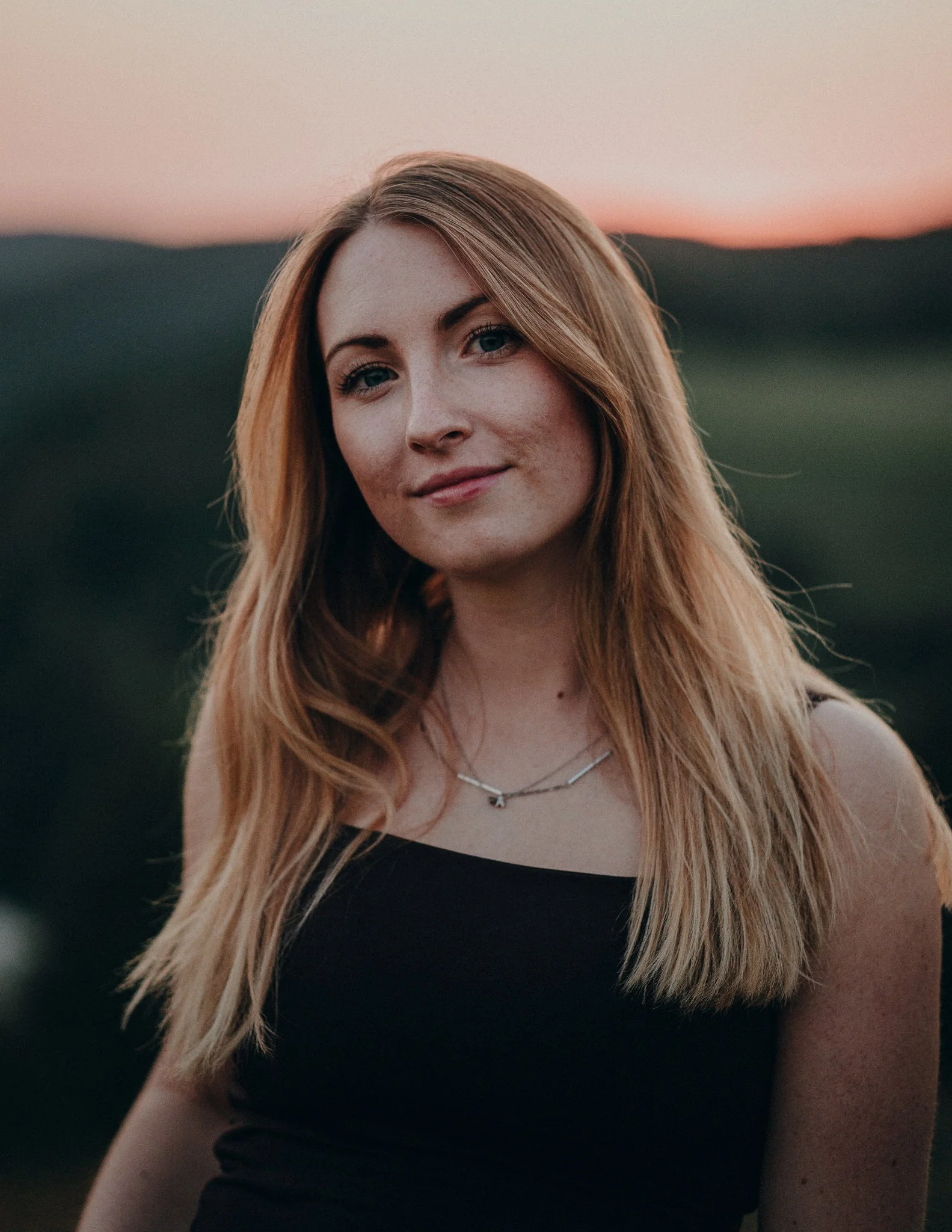 Junge Frau mit langem, lockigem roten Haar, trägt schwarze Oberteil und silberne Halskette, im Freien bei Sonnenuntergang.