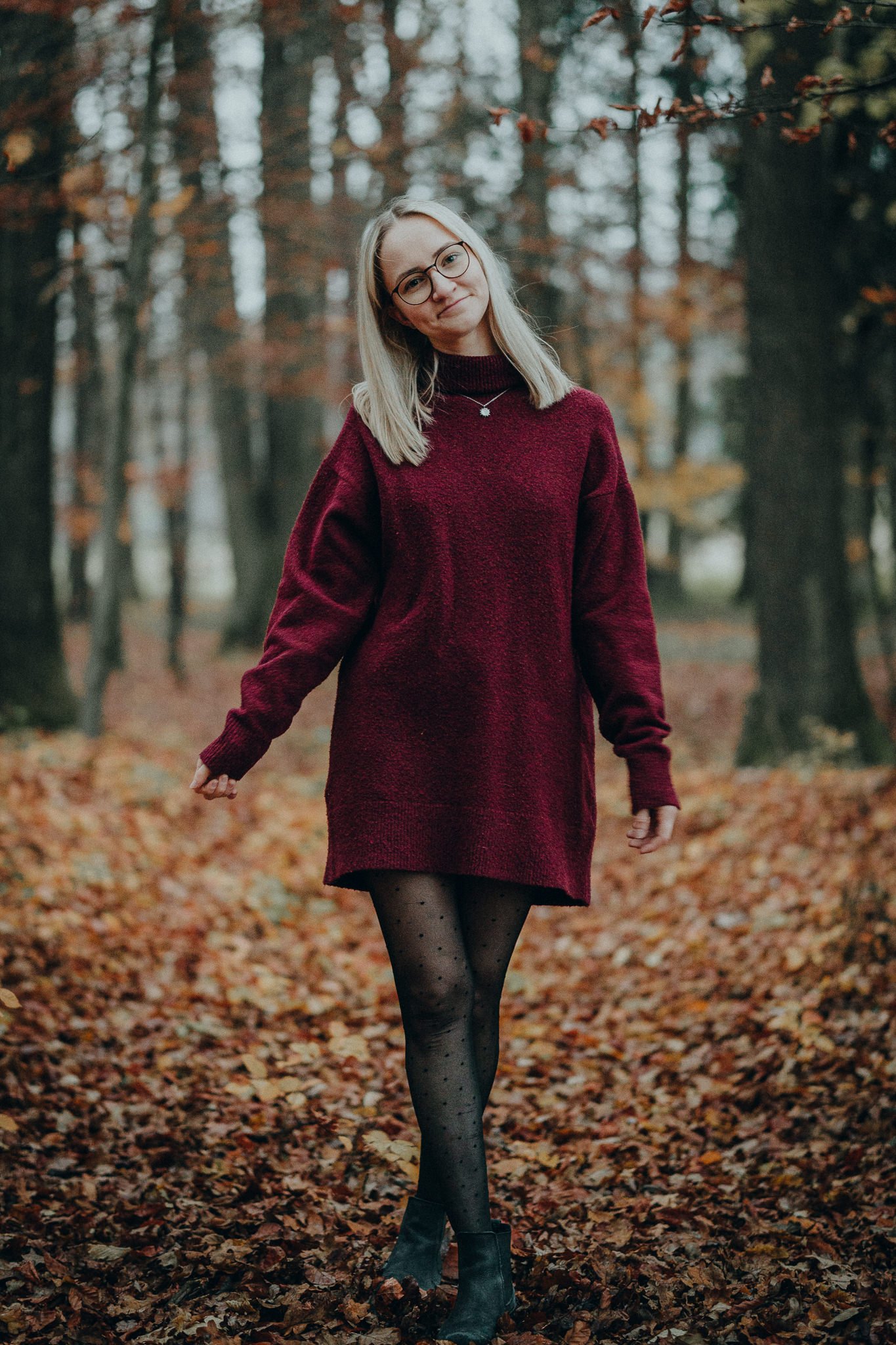 Junge Frau in rotem Pullover, Schwarze Strumpfhose mit Punkten, schwarzen Stiefeln, steht im Herbstlaub im Wald.