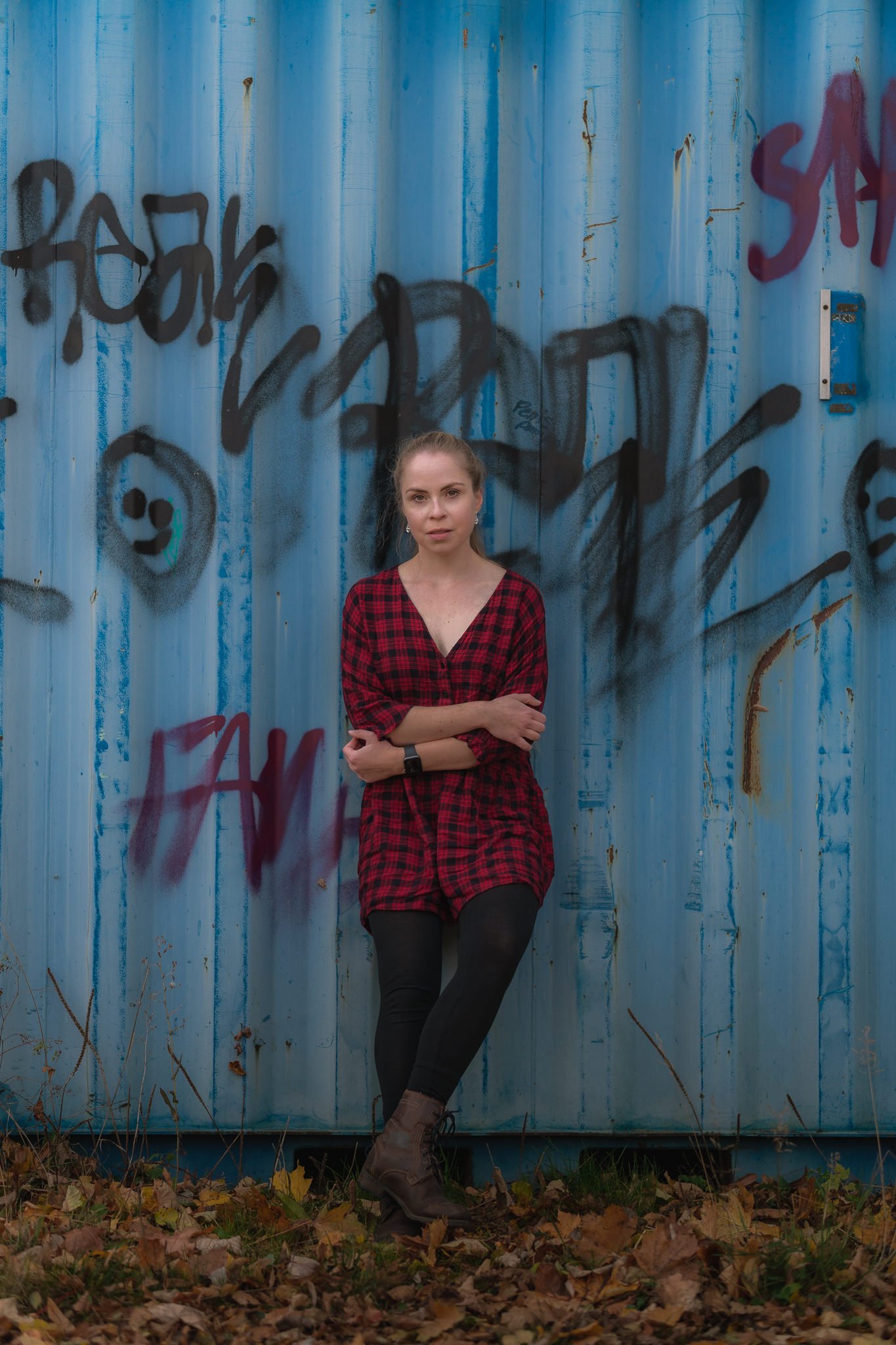 Eine junge Frau in rotem Flanellhemd und schwarzen Leggings lehnt an einer mit Graffiti besprayten blauen Metallwand. Sie trägt braune Stiefel und hat lange blonde Haare. Der Boden ist mit Herbstlaub bedeckt.