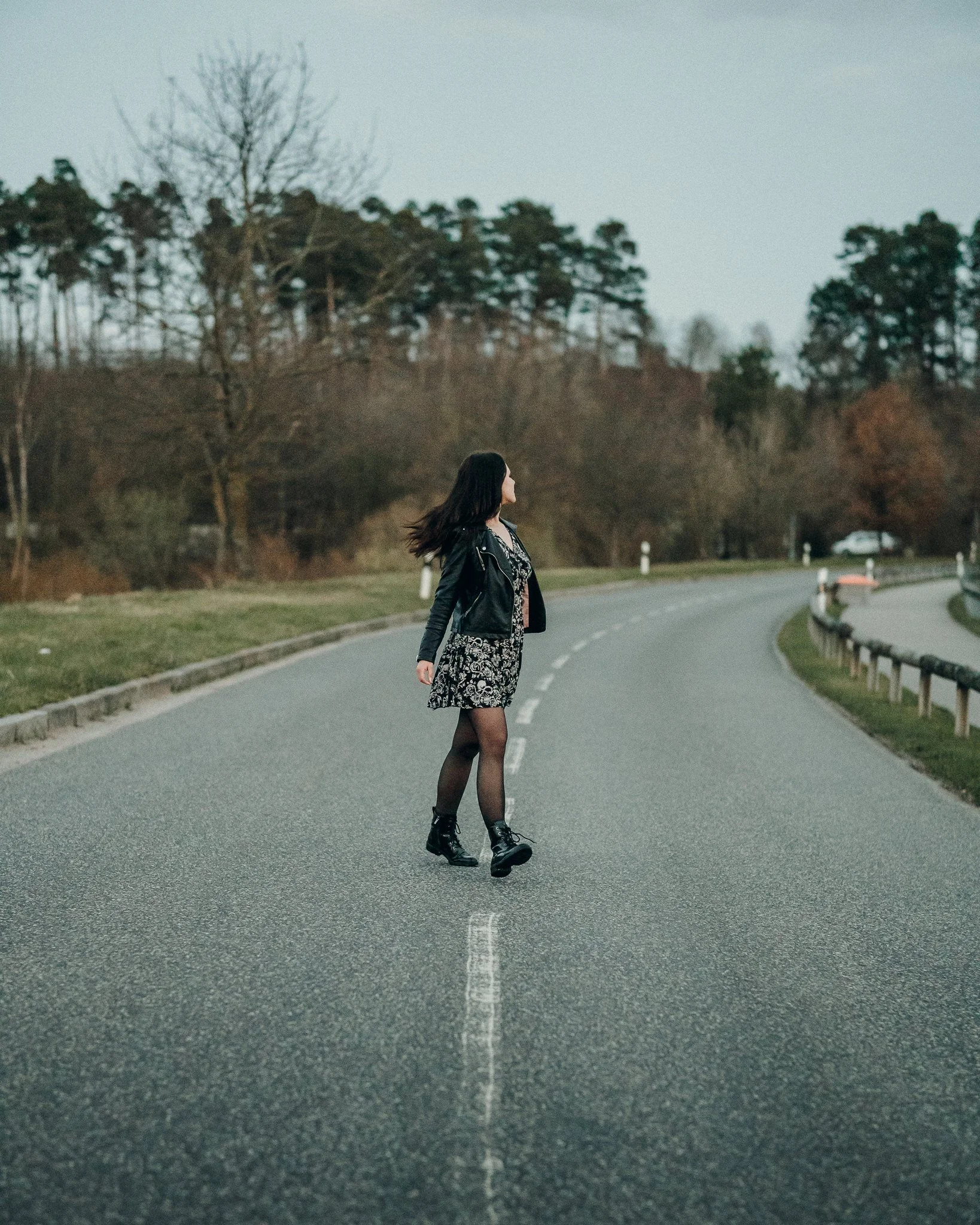 Frau steht in der Mitte einer leeren Straße, umgeben von Bäumen, trägt schwarze Stiefel, einen schwarzen Rock mit Muster, eine schwarze Lederjacke und hat lange dunkle Haare.