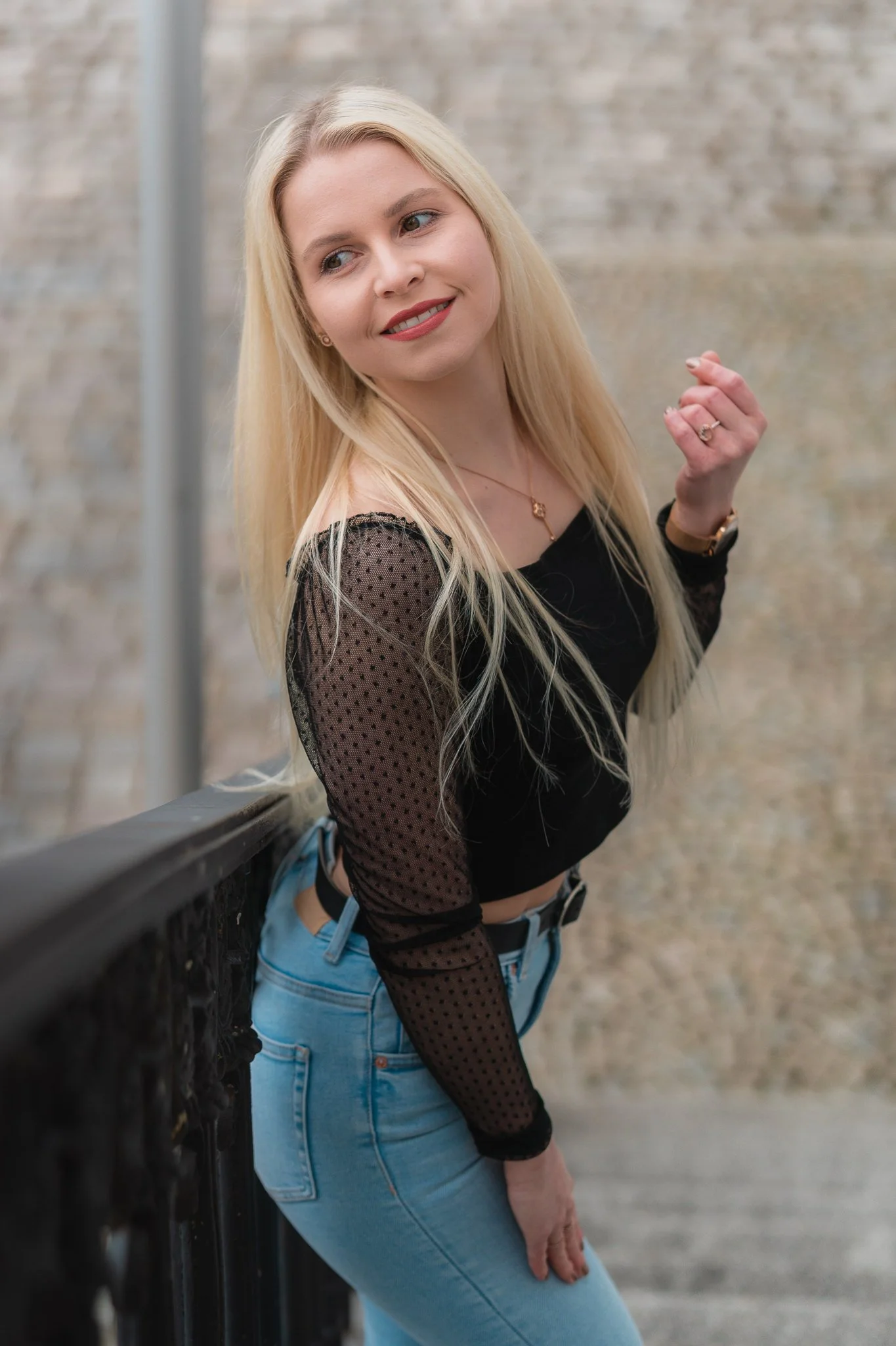 Junge Frau mit blonden Haaren, schwarze transparente Bluse und blaue Jeans, steht an einer Brüstung vor einer Backsteinwand, posiert und lächelt.